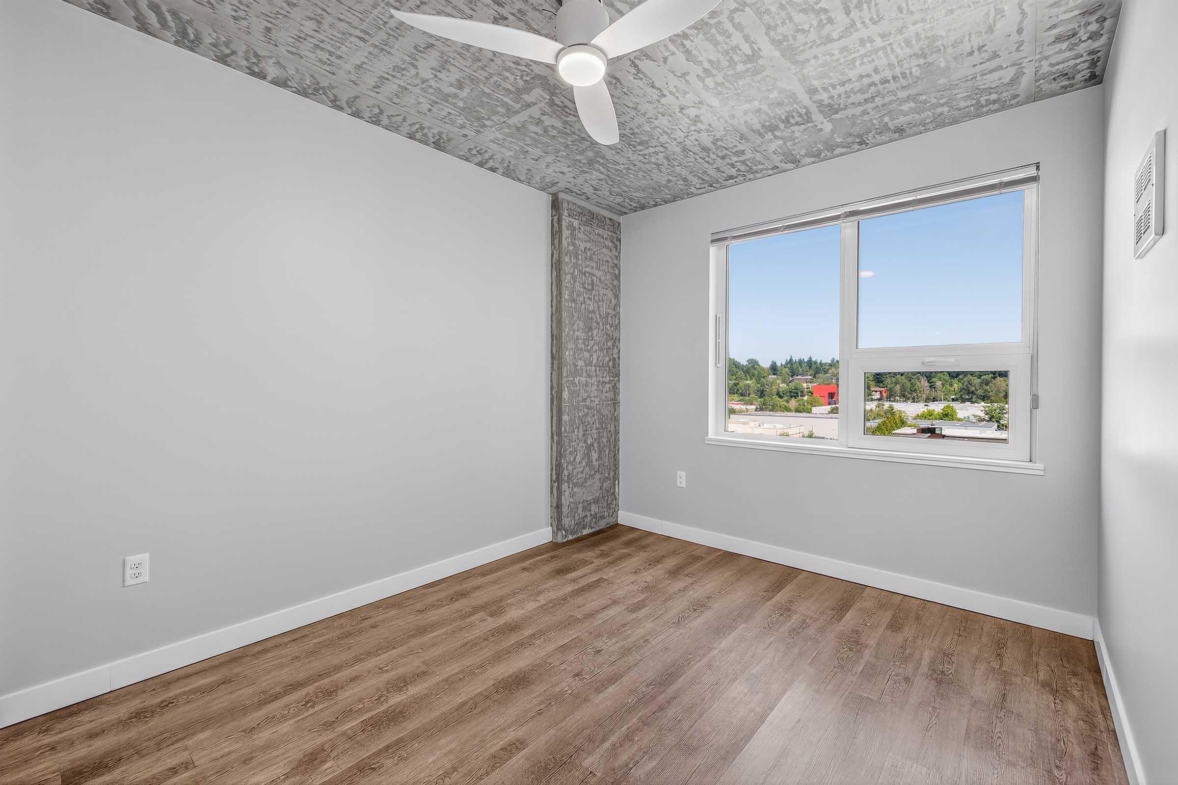 A well-lit empty room with a gray ceiling and wall accents. The floor is wood-like, and there is a large window providing natural light. The walls are painted light gray, offering a modern and minimalist aesthetic. Ideal for various uses such as a bedroom or office space.