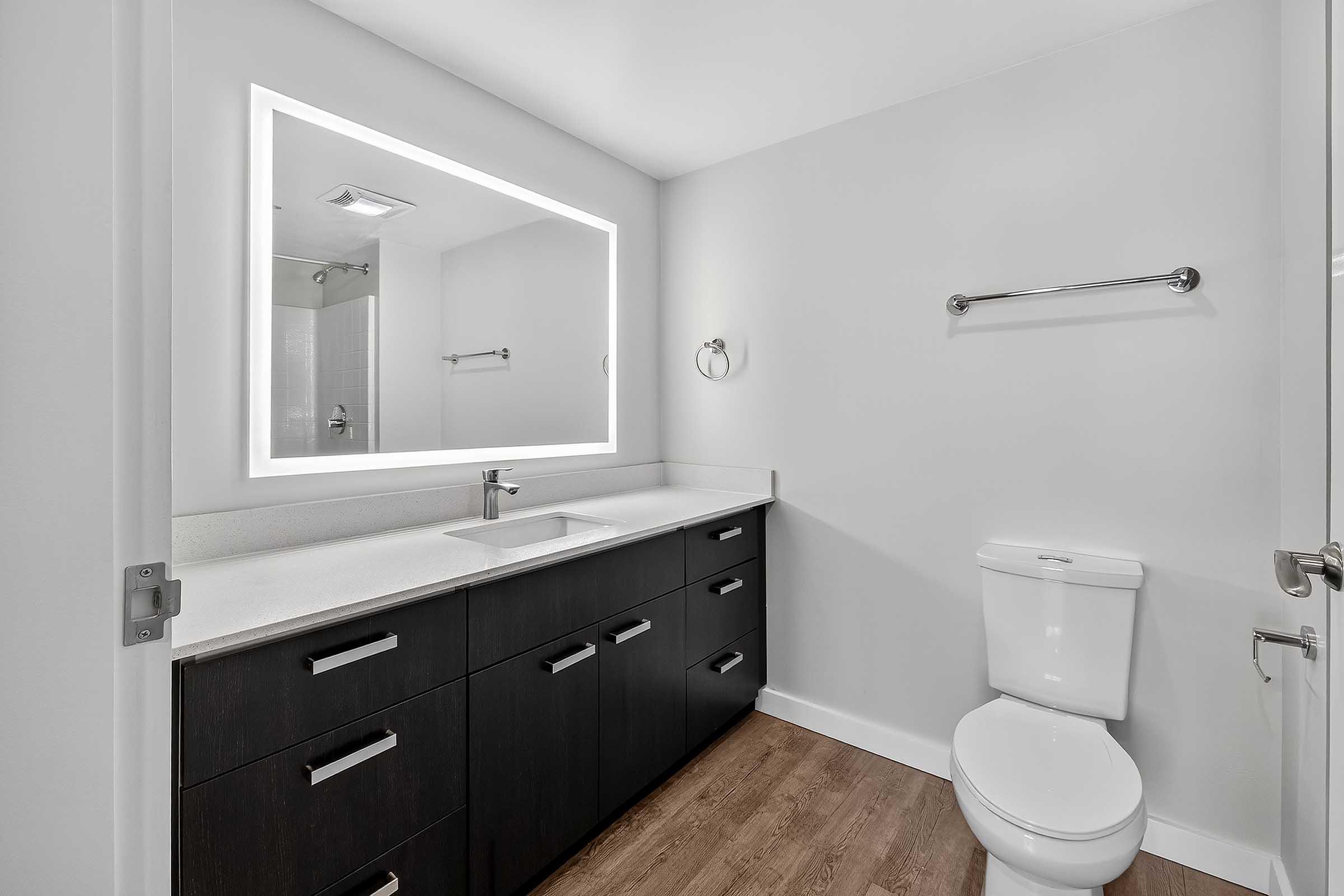 Modern bathroom featuring a sleek design with a white countertop, dark cabinetry, and a backlit mirror. The space includes a toilet and a towel rack, complemented by neutral-colored walls and light wooden flooring. Spacious and well-lit, offering a clean aesthetic.