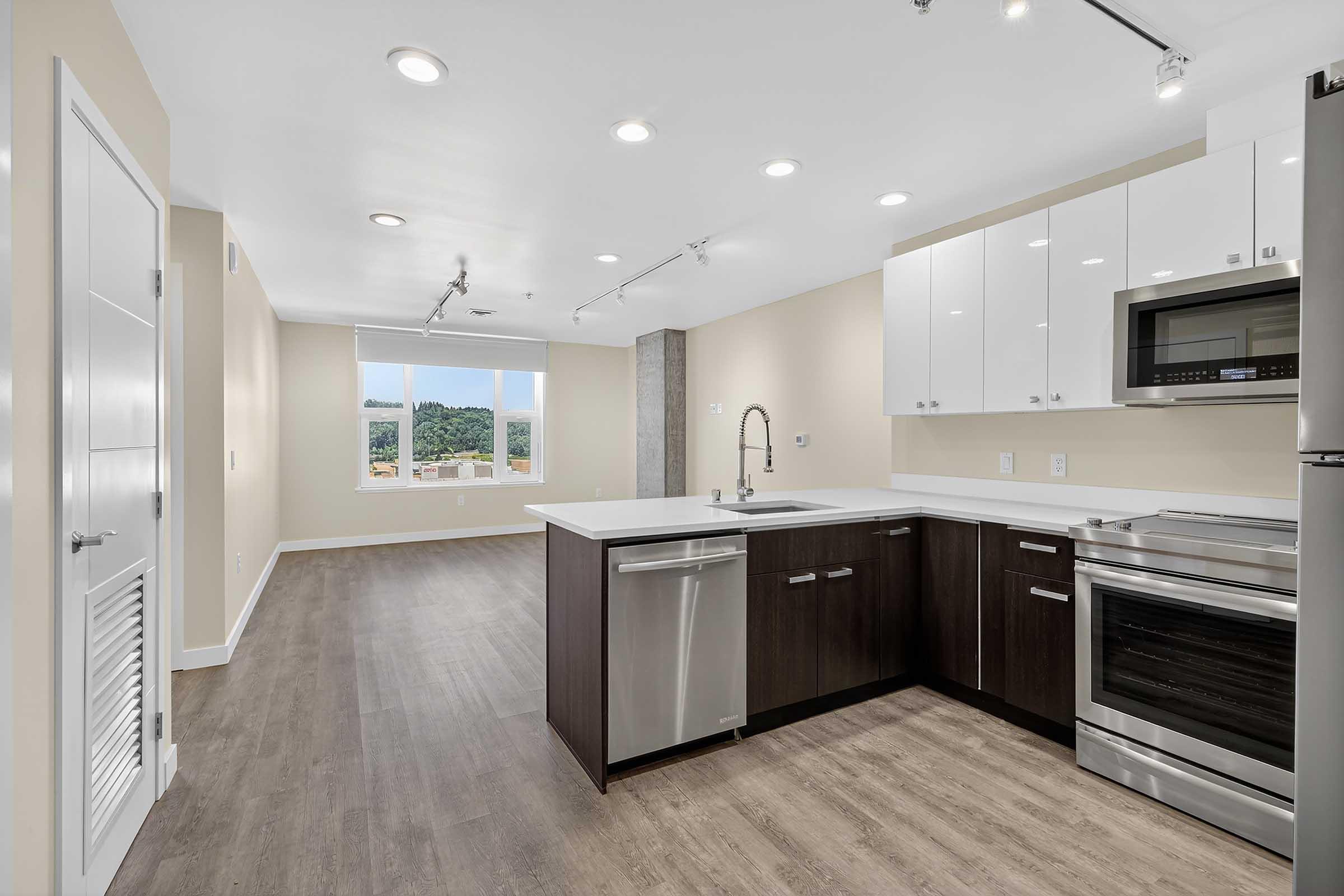 Modern kitchen featuring a spacious layout with stainless steel appliances, including a microwave and dishwasher. The cabinets are a mix of white and dark wood, and the countertops are light-colored. Bright, recessed lighting and a large window provide natural light, with a view of greenery outside.
