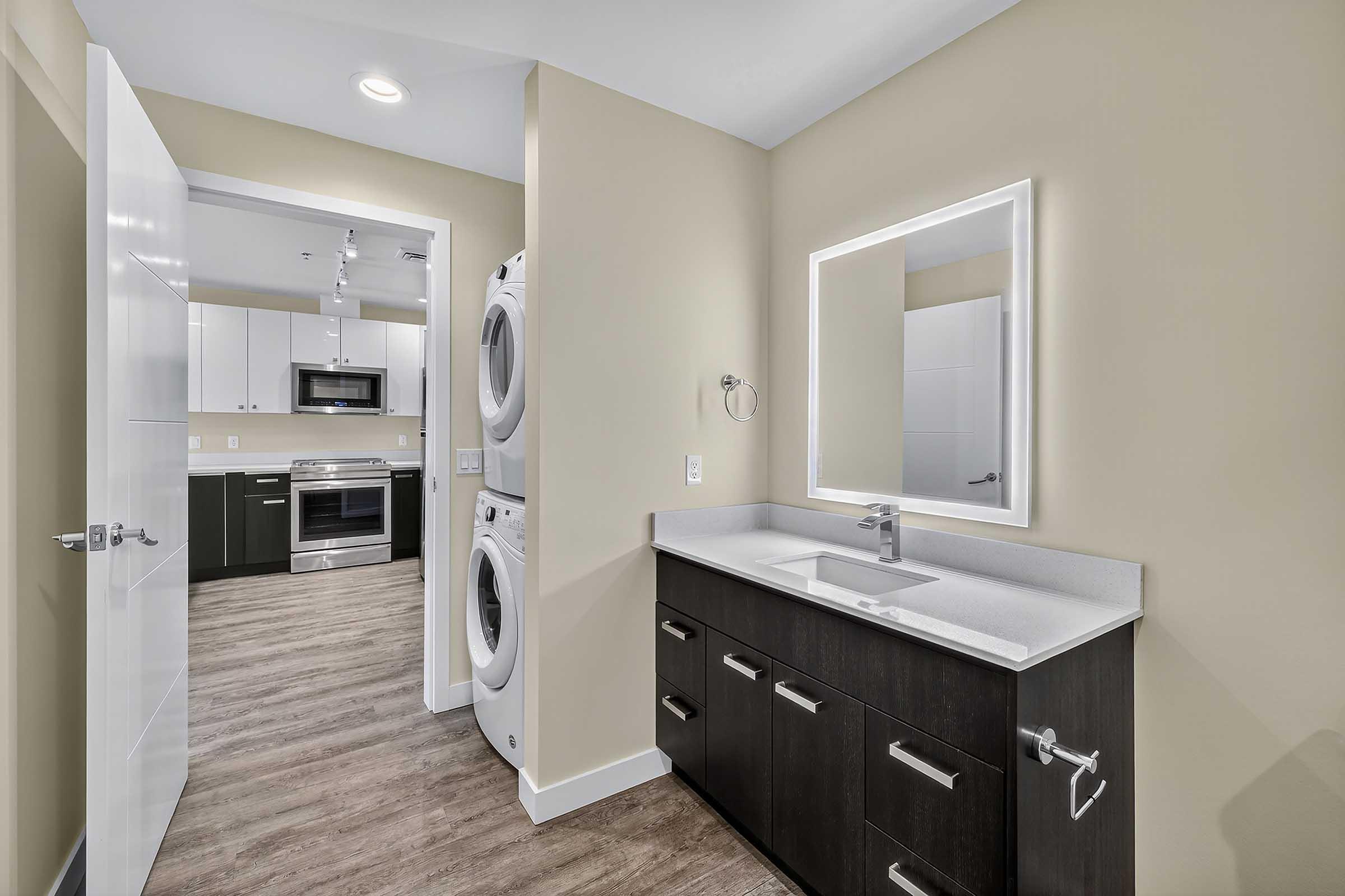 Contemporary laundry room with stacked washer and dryer, featuring a sleek vanity with a sink and modern faucet. The space is well-lit with a mirror and stylish wall-mounted accessories, complemented by light-colored walls and a wood-like floor. A kitchen area is visible through an open doorway.
