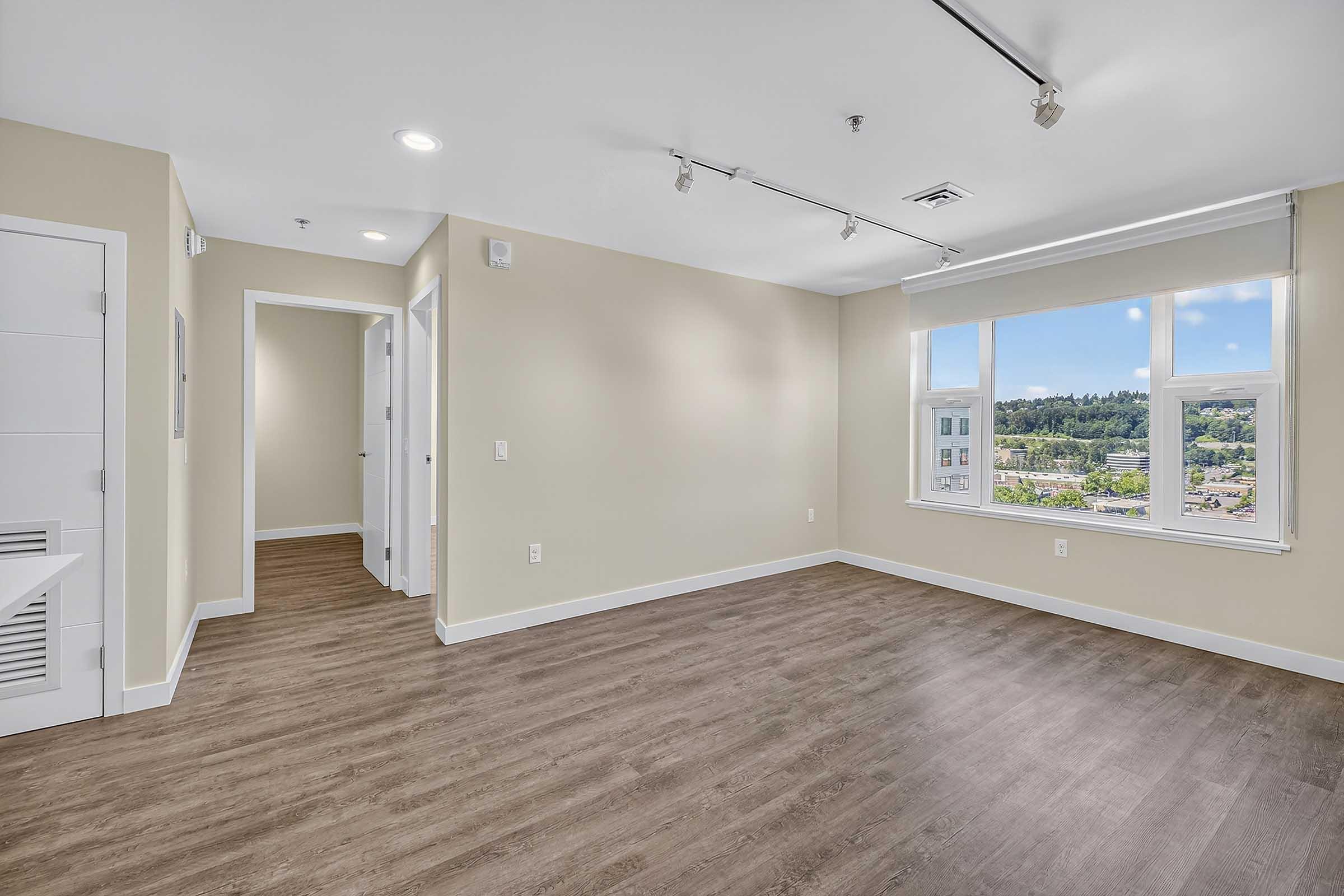Bright and airy empty room with beige walls and large windows allowing natural light. Features include track lighting and modern laminate flooring. A doorway leads to another room and a closet is visible on the left. The scenic view outside showcases greenery and buildings in the distance.