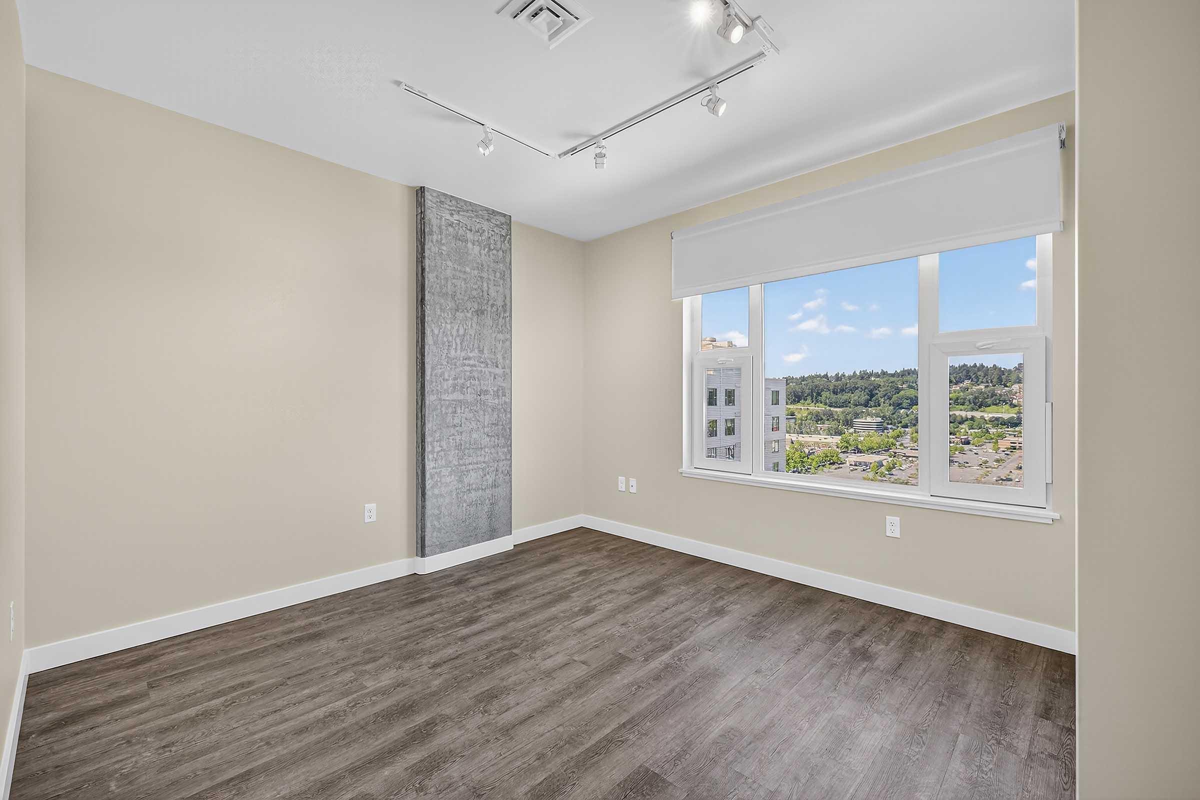 A modern, empty room featuring light beige walls, large windows with a view, and attractive wood-like flooring. The room is illuminated by track lighting and has a textured concrete accent wall, adding a contemporary touch to the space.