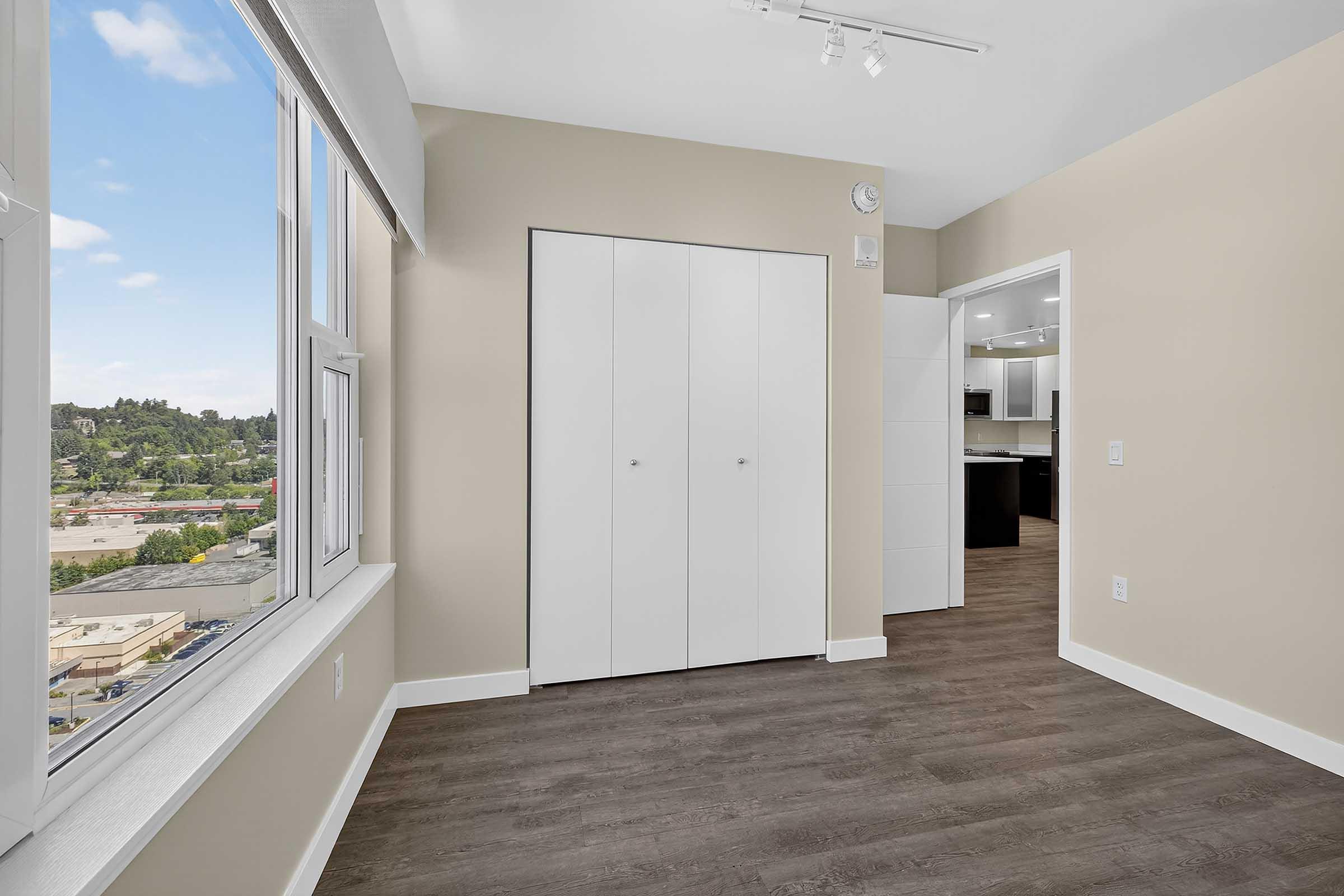 A well-lit room with light-colored walls and a large window offering a view of greenery and buildings outside. The floor is covered with dark laminate. There is a white closet with sliding doors on the wall, and a doorway leads to a kitchen area visible in the background.