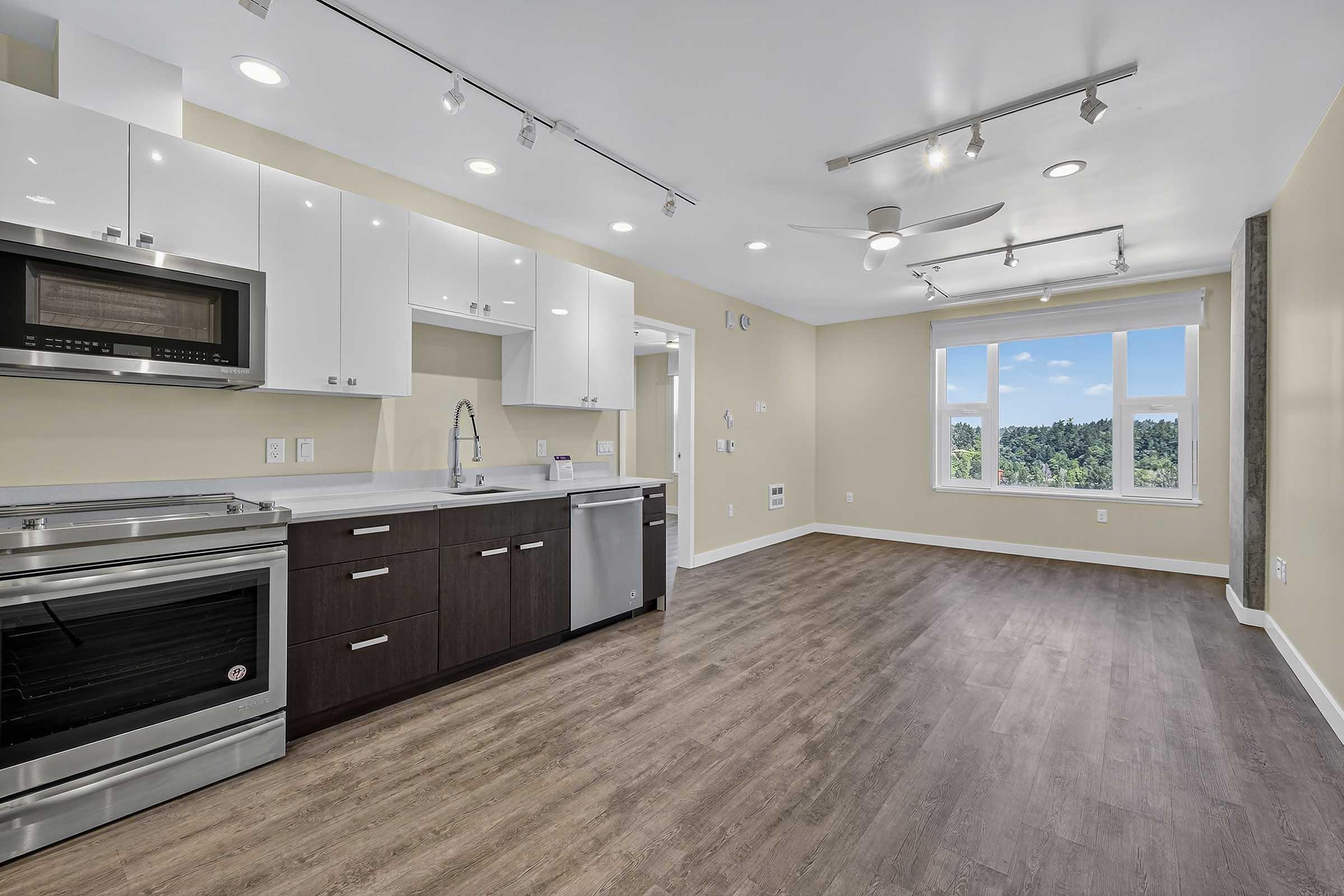 Modern kitchen and living area featuring sleek cabinetry, stainless steel appliances, and an island. Large windows provide natural light and a view of the outdoors. The flooring is a warm hardwood, complementing the neutral wall colors and contemporary lighting fixtures.