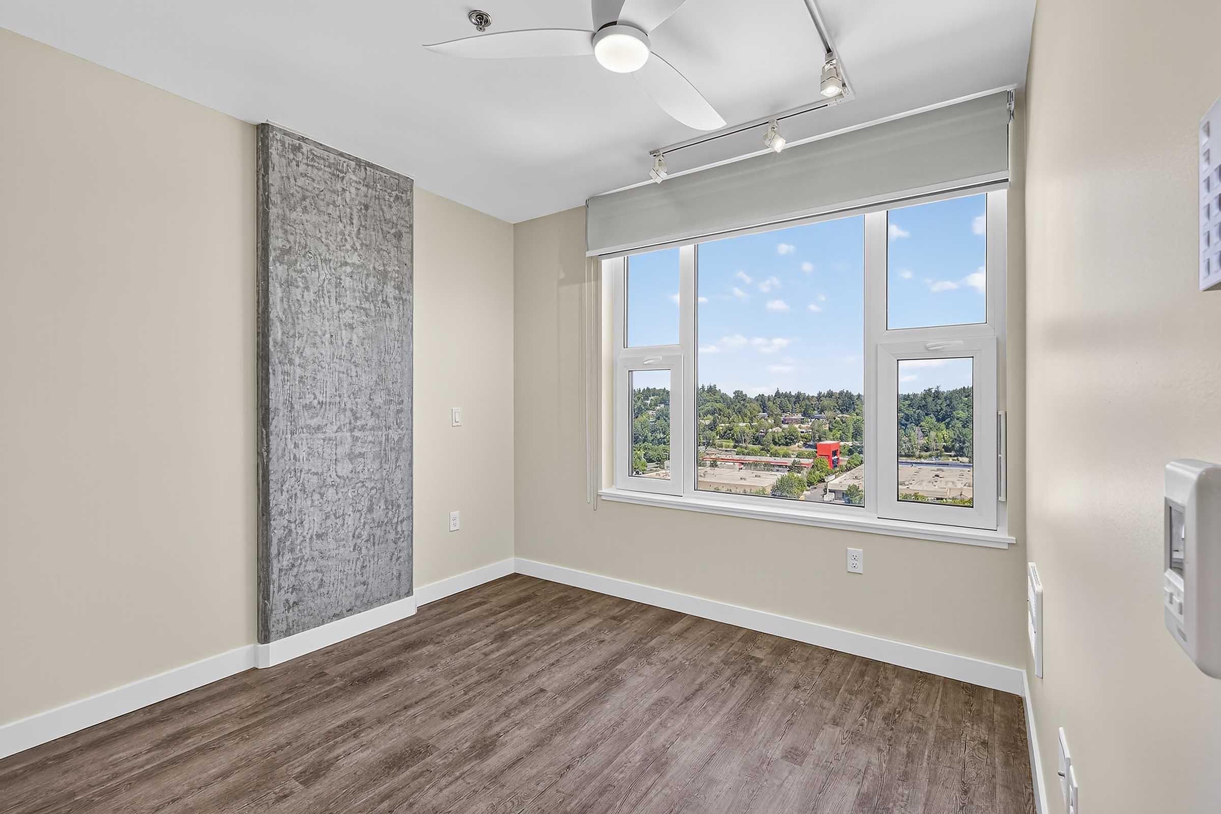 A well-lit, empty room with light beige walls and dark wood laminate flooring. A large window offers a view of greenery outside, while a ceiling fan is installed. A gray textured wall panel is visible on one side, creating visual interest. The overall ambiance is modern and inviting.