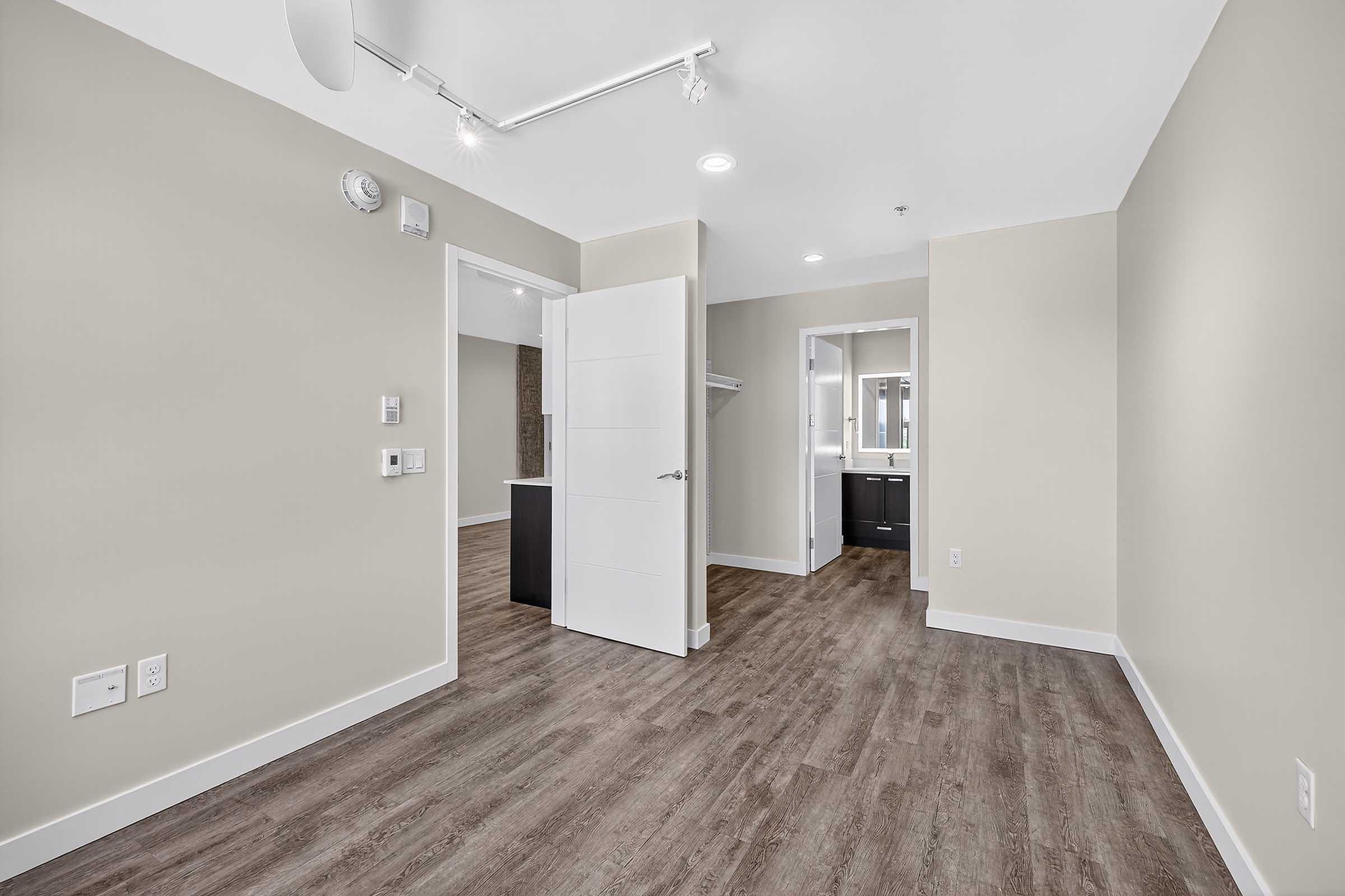 A well-lit, modern room featuring hardwood-style flooring, light beige walls, a white door, and an open doorway leading to another space. Wall-mounted fixtures and two adjacent entryways provide access to different areas, creating a spacious and inviting atmosphere.