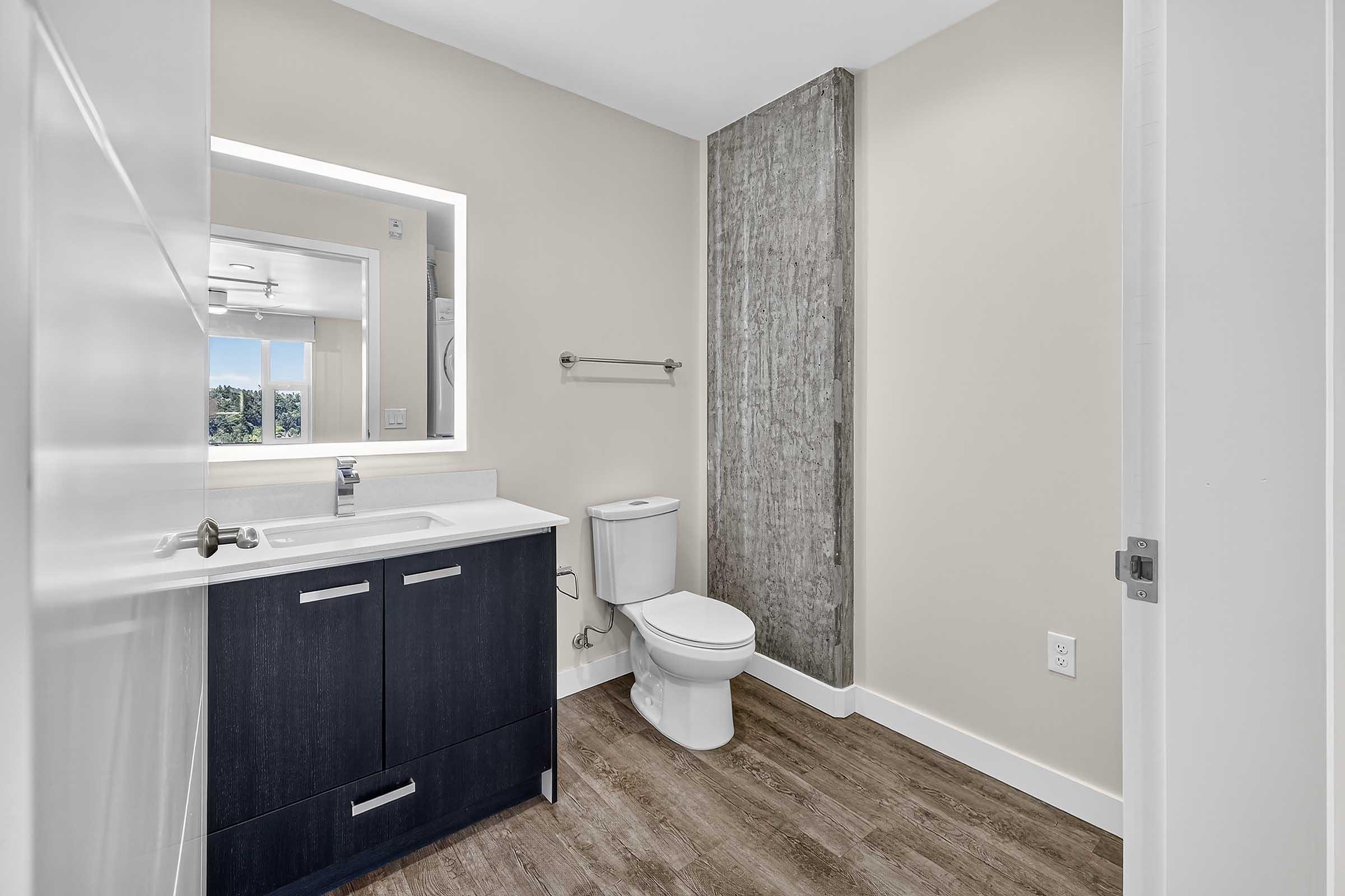 A modern bathroom featuring a dark wood vanity with a white countertop, a mirror with built-in lighting, a toilet, and a textured concrete wall. The floor is adorned with light wood-style tiles, and there is a window providing natural light. The overall design is sleek and contemporary.