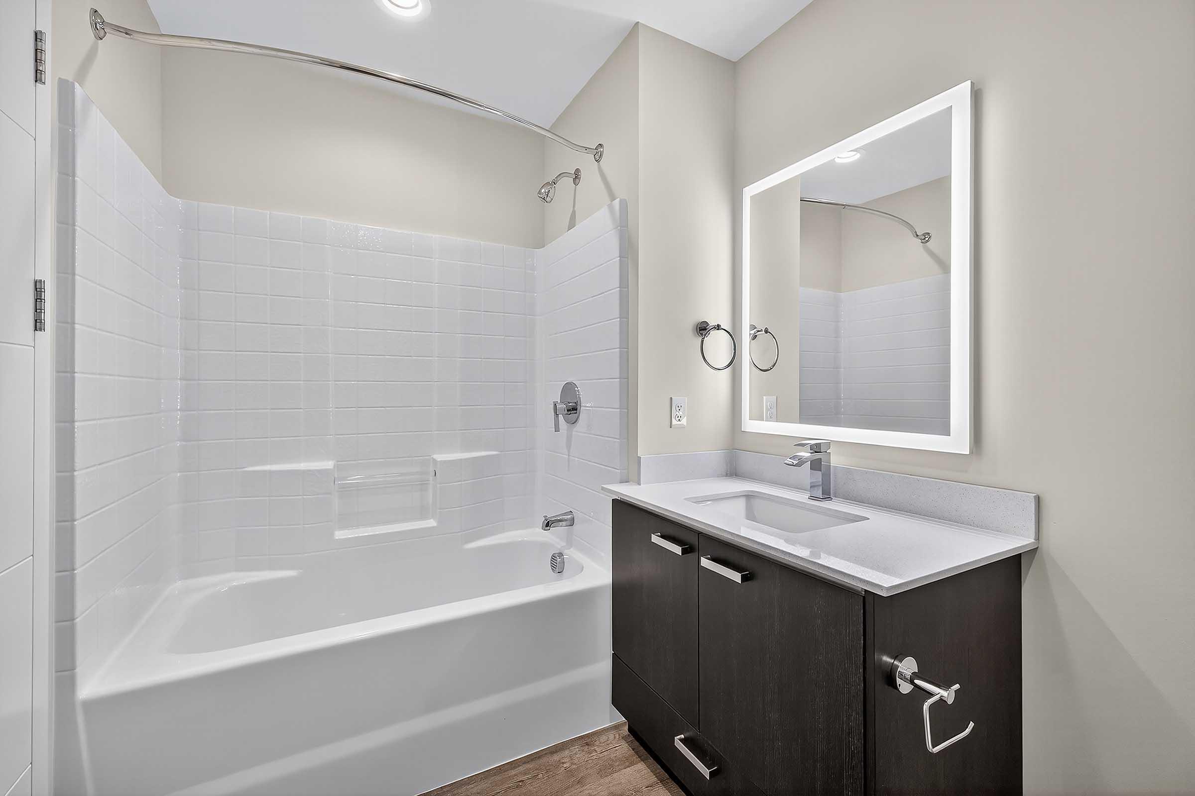 A modern bathroom featuring a white bathtub with a curved shower rod and white tiled walls. There is a sleek vanity with a sink and a large mirror above it. The walls are painted a soft neutral color, and the floor is made of light-colored wood.