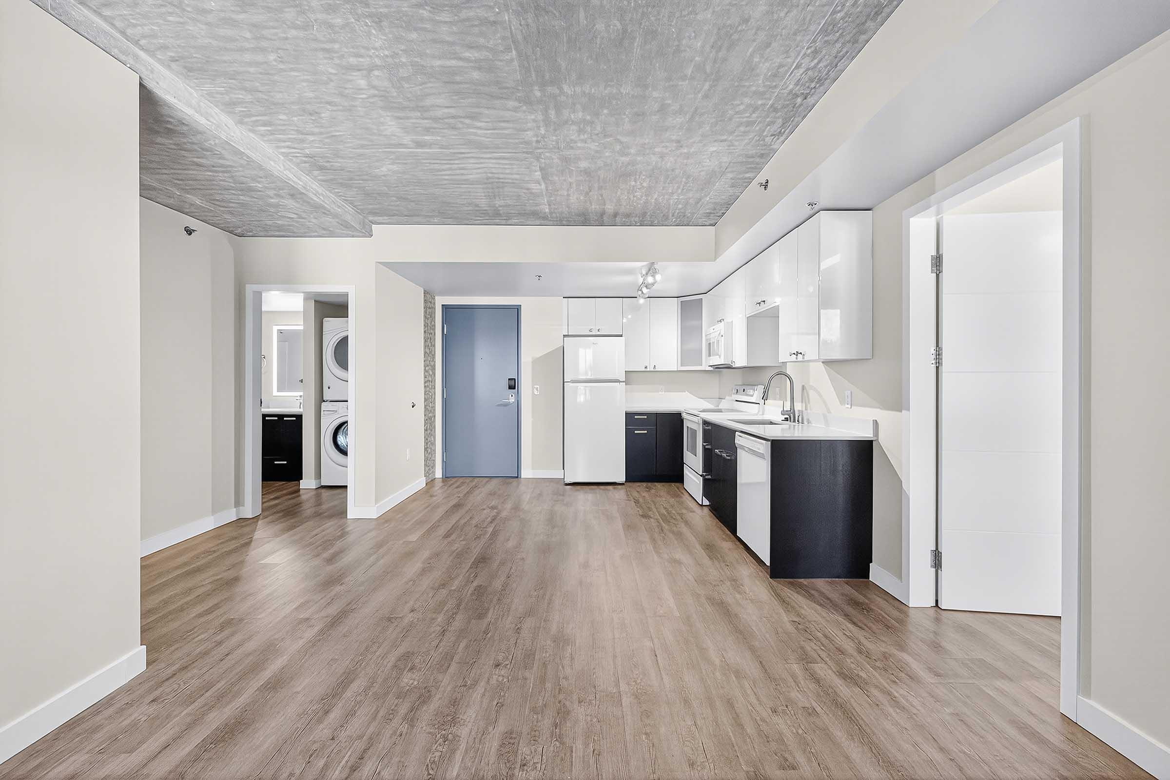Modern open-concept kitchen and living area featuring sleek white cabinets, stainless steel appliances, and a neutral color palette. Large window allows natural light to brighten the space. Laundry area visible through a door on the left. The floor is finished with light wood laminate, enhancing the contemporary feel.