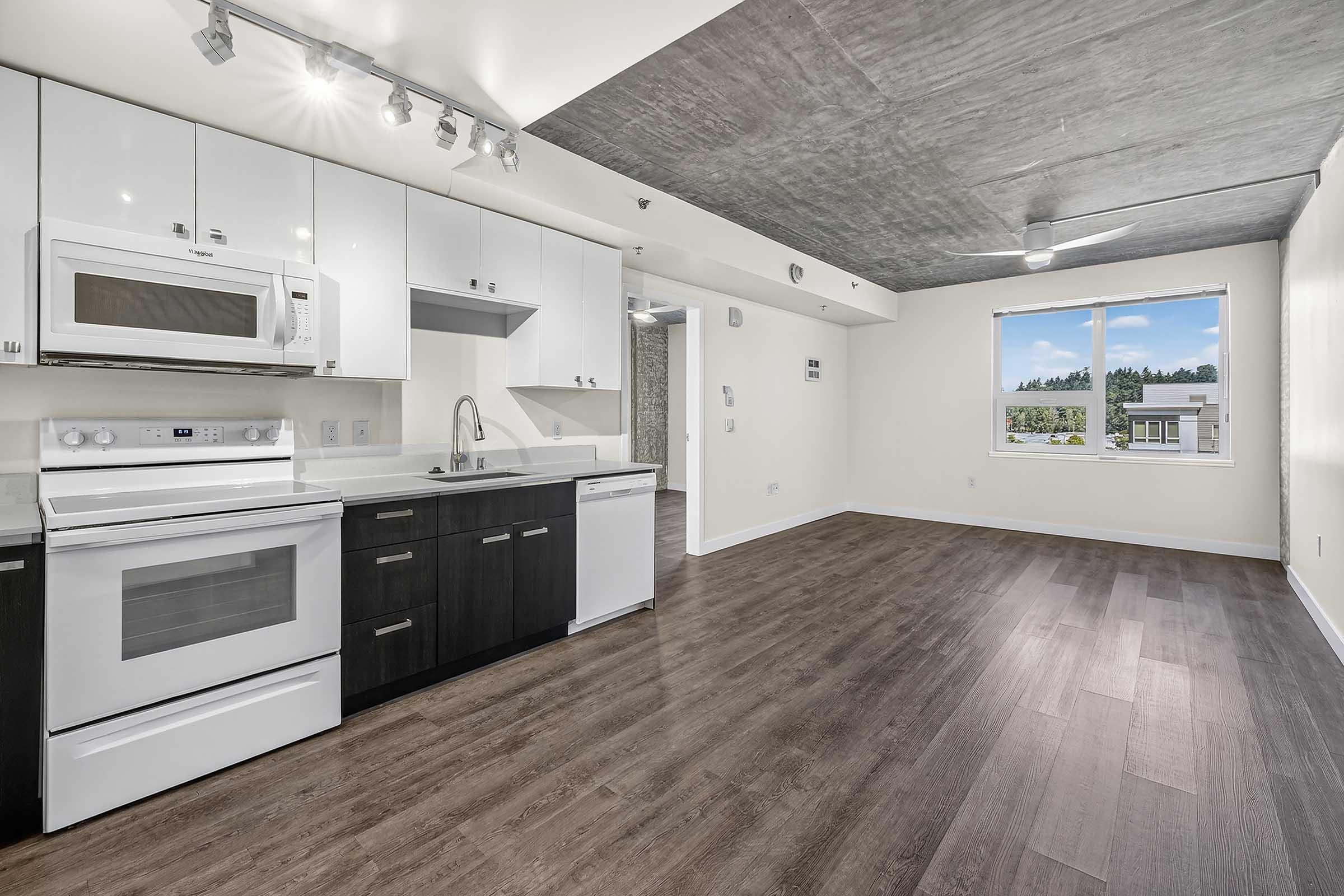 Modern kitchen featuring white and dark cabinetry, stainless steel appliances including a microwave and dishwasher, and a spacious layout. The room has hardwood flooring and large windows that provide natural light, with a view of the outdoors. Ceiling with exposed concrete adds a contemporary touch.