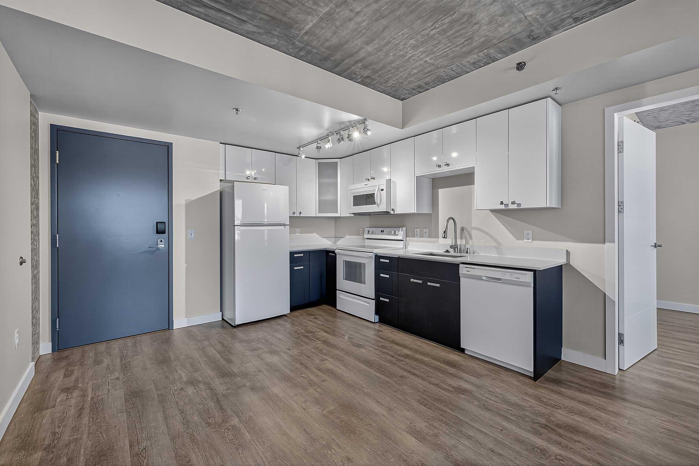 Modern kitchen in an apartment featuring white and dark cabinetry, stainless steel appliances including a refrigerator, oven, and dishwasher, a double sink, and wood-style flooring. The space has neutral walls and a contemporary ceiling design.