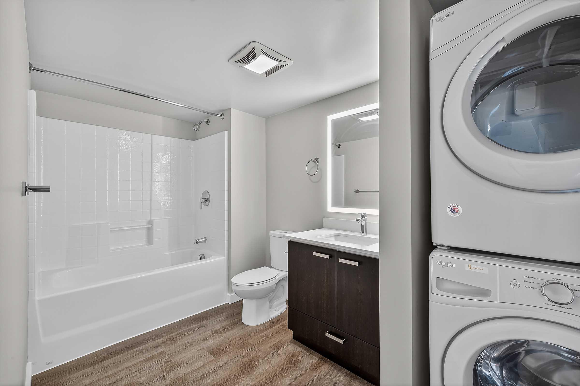 A modern bathroom featuring a bathtub with a shower, a toilet, a sink with a cabinet, and a stacked washer and dryer unit. The walls are painted in a light color, and there is a mirror with built-in lighting above the sink, with a wood-like floor completing the contemporary design.
