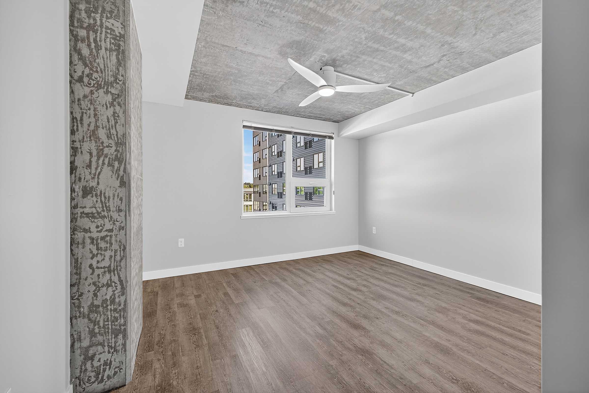 Empty modern room with a concrete ceiling and a large window allowing natural light. A ceiling fan is installed, and the flooring is wood. The walls are painted in a light color, creating a spacious and airy feel.