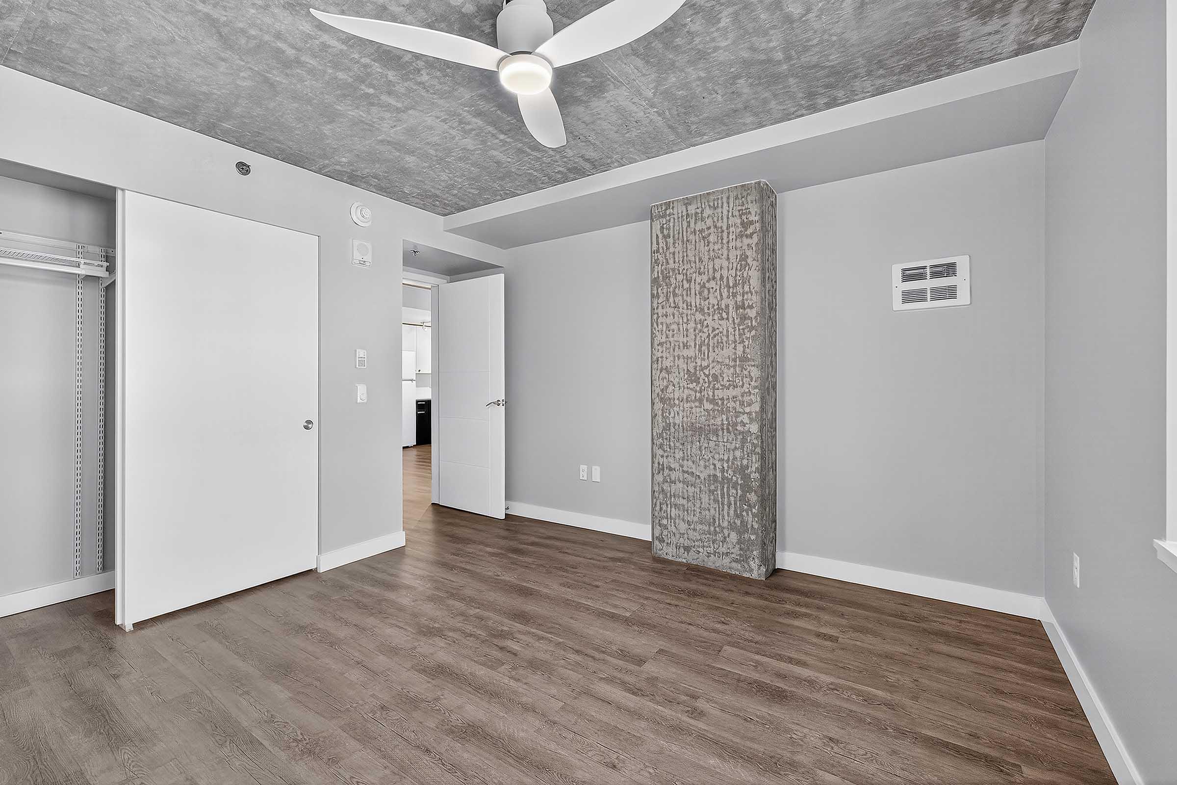 A modern, empty room featuring grey walls and a textured ceiling. The space has a ceiling fan, a closet with sliding doors, and a tall, textured column along one wall. The floor is made of wood or laminate, giving a warm appearance. A small vent is visible on the wall.