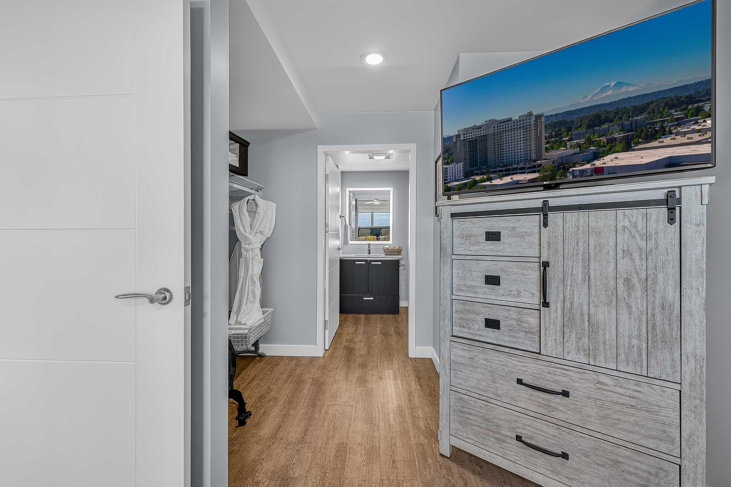 A modern interior space featuring a hallway leading to a bathroom and another room. The hallway is framed by a white door and has wooden flooring. A dresser with multiple drawers is visible on the left, and a large TV mounted above it displays an outdoor view, including a mountain in the background.