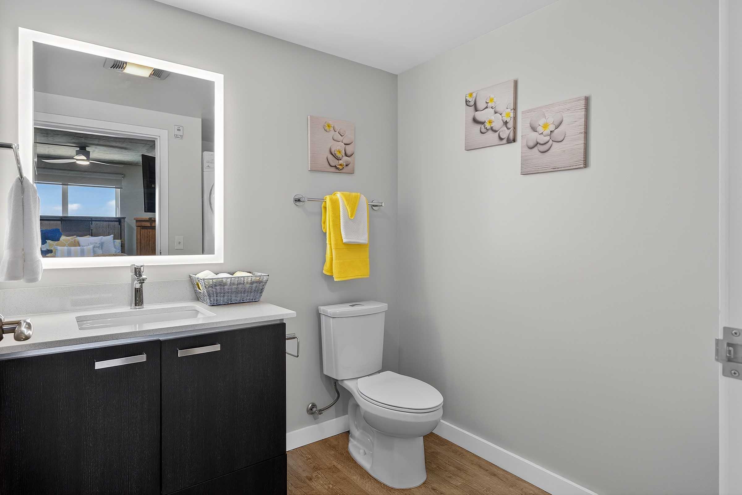 A modern bathroom with white walls and a mirror featuring LED lights. It includes a dark wood vanity with a sink, a neatly arranged towel in yellow, and a decorative basket. The toilet is positioned beside the vanity, and there are three flower-themed wall art pieces above the towel rack. Natural light enters from a nearby window.
