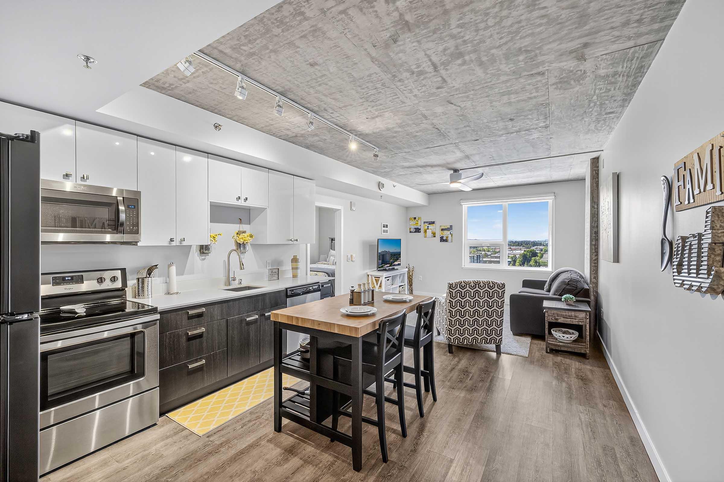 Modern kitchen and living area featuring stainless steel appliances, a breakfast bar with stools, and natural light from a large window. The decor includes a gray couch, a patterned chair, and wall art. The ceiling has an industrial finish, and a yellow rug adds a pop of color to the space.