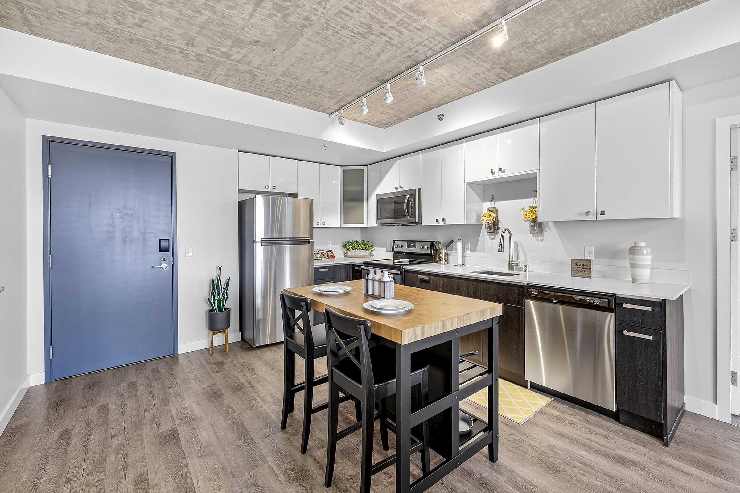 Modern kitchen with a central wooden island featuring two place settings, white cabinets, stainless steel appliances, and a sleek design. The space has a polished concrete ceiling and wooden flooring, creating a contemporary feel. A blue door adds a pop of color to the minimalist decor.