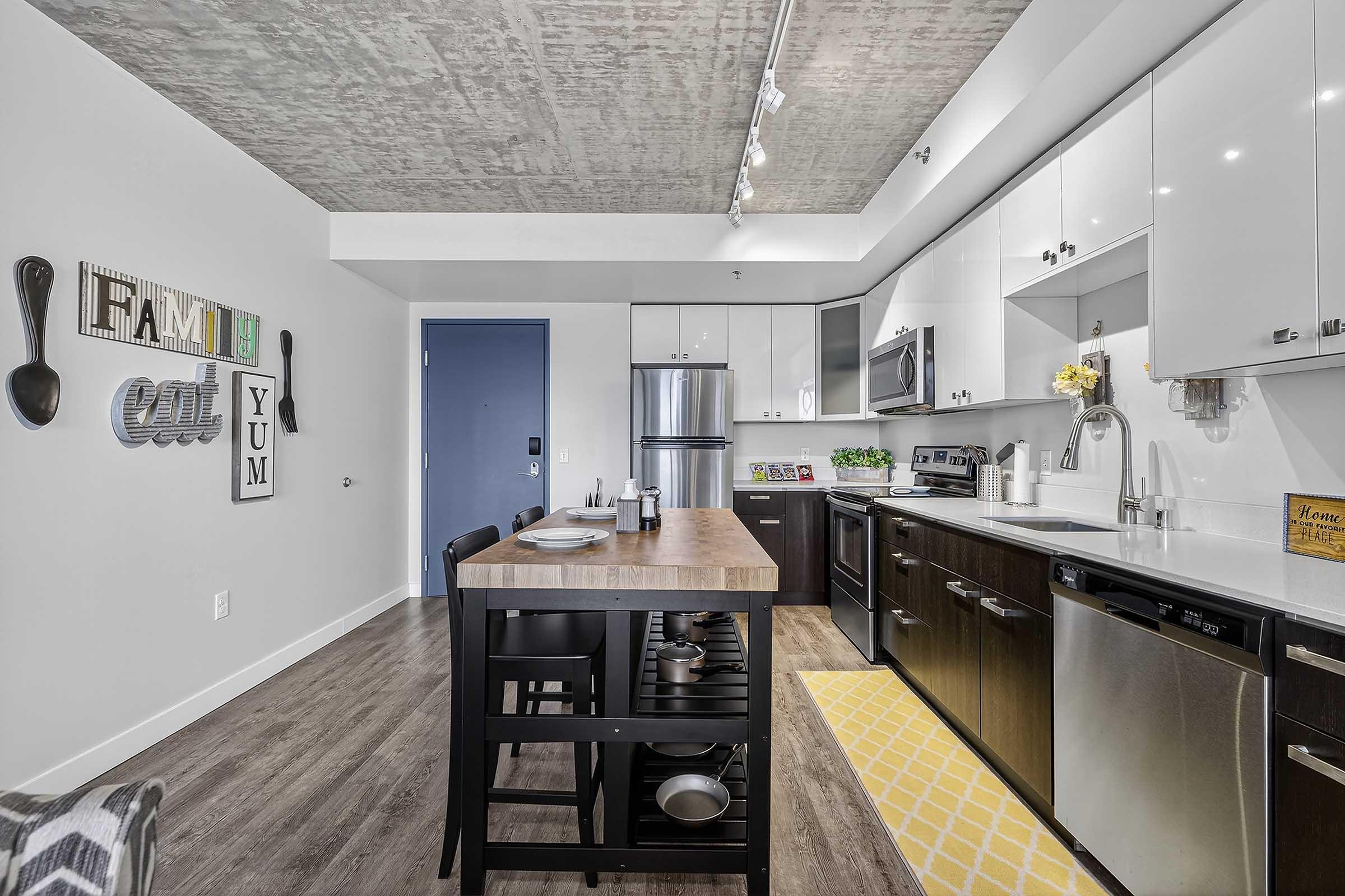 Modern kitchen featuring a concrete ceiling, sleek cabinetry, stainless steel appliances, and a central island with seating. Decor includes wall art with words like "Family" and "Yum." A bright yellow rug adds a pop of color to the wooden floor. Natural light enhances the spacious, contemporary feel.