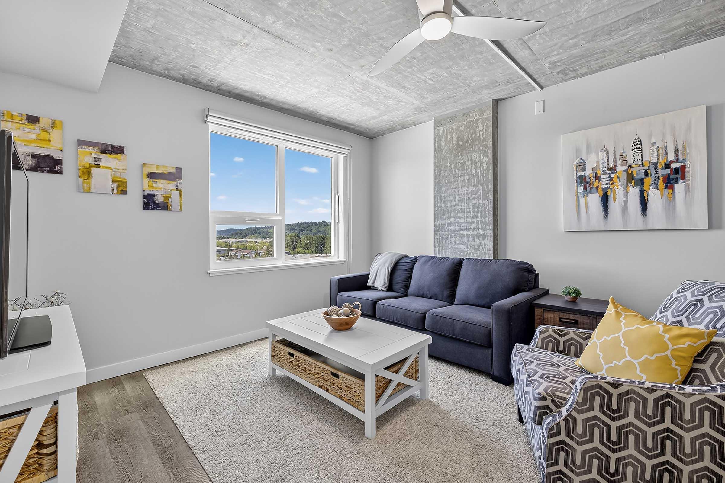 A modern living room featuring a navy blue sofa and an accent chair with a geometric pattern. A white coffee table is centered on a soft rug, with decorative items on top. Large windows offer a scenic view, and abstract wall art adds color to the gray walls and ceiling.