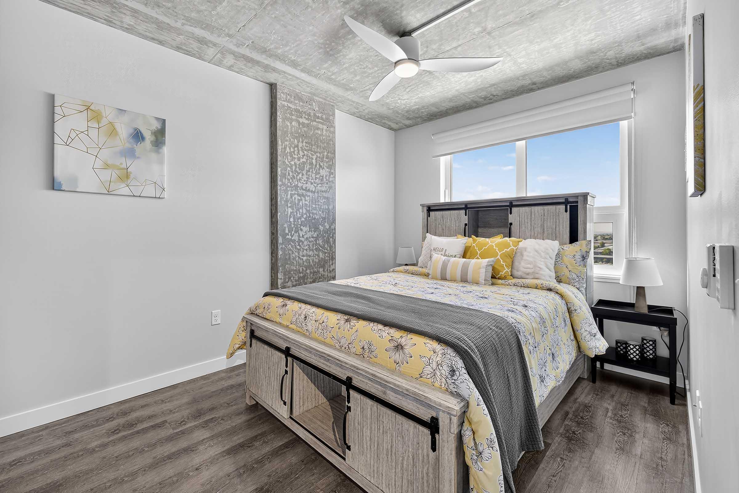 A stylish bedroom featuring a queen-sized bed with a yellow and gray floral comforter, decorative pillows, and a wooden bed frame. A modern ceiling fan hangs above, and a window allows natural light to flood the space. There’s a textured wall, minimalist artwork, and a nightstand with a lamp. The floor is wood-look laminate.