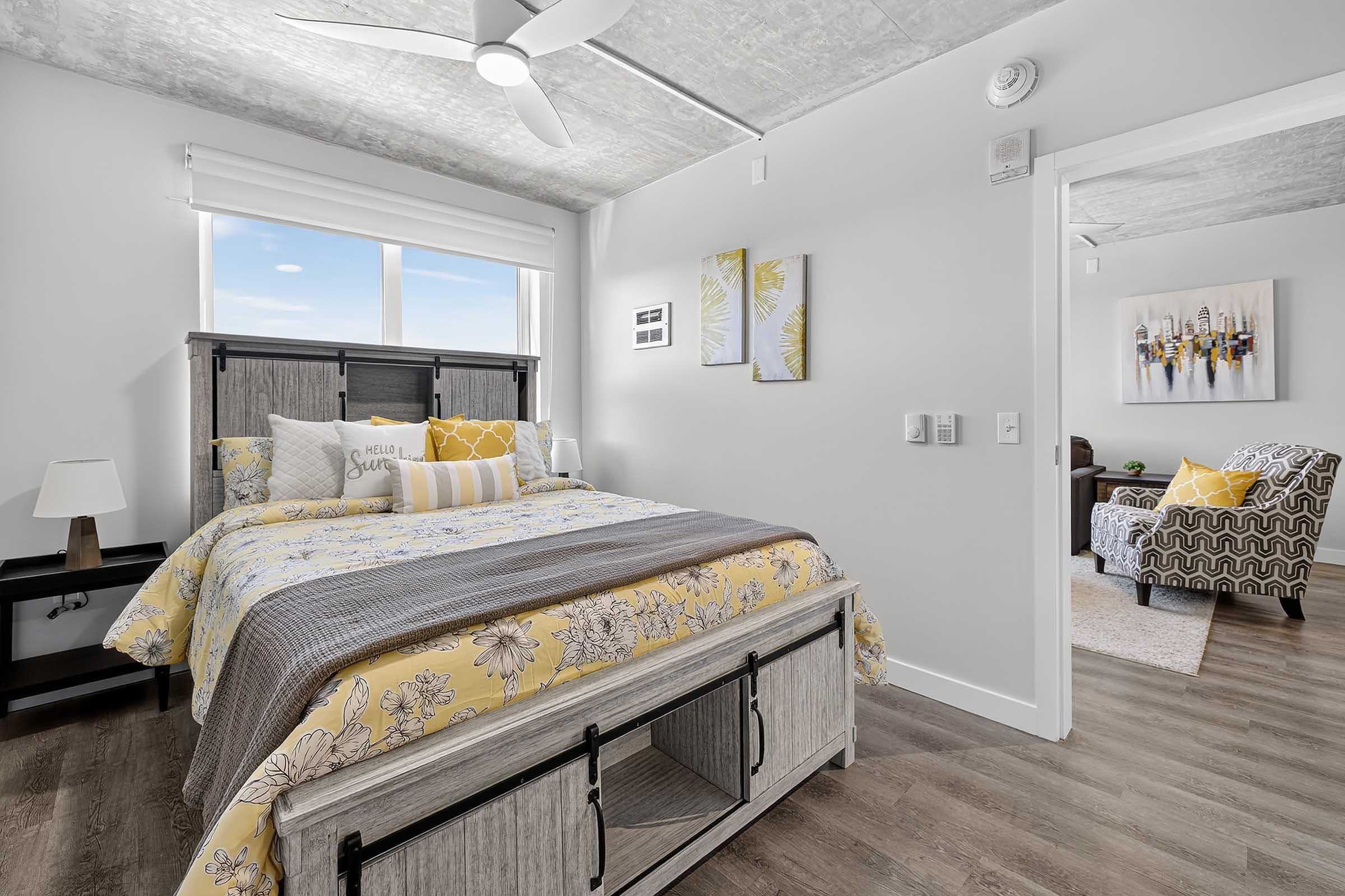 A bright, modern bedroom featuring a queen-sized bed with yellow floral bedding, a bedside table with a lamp, and a large window letting in natural light. The decor includes two framed pictures on the wall. In the background, there's a cozy sitting area with a patterned armchair.