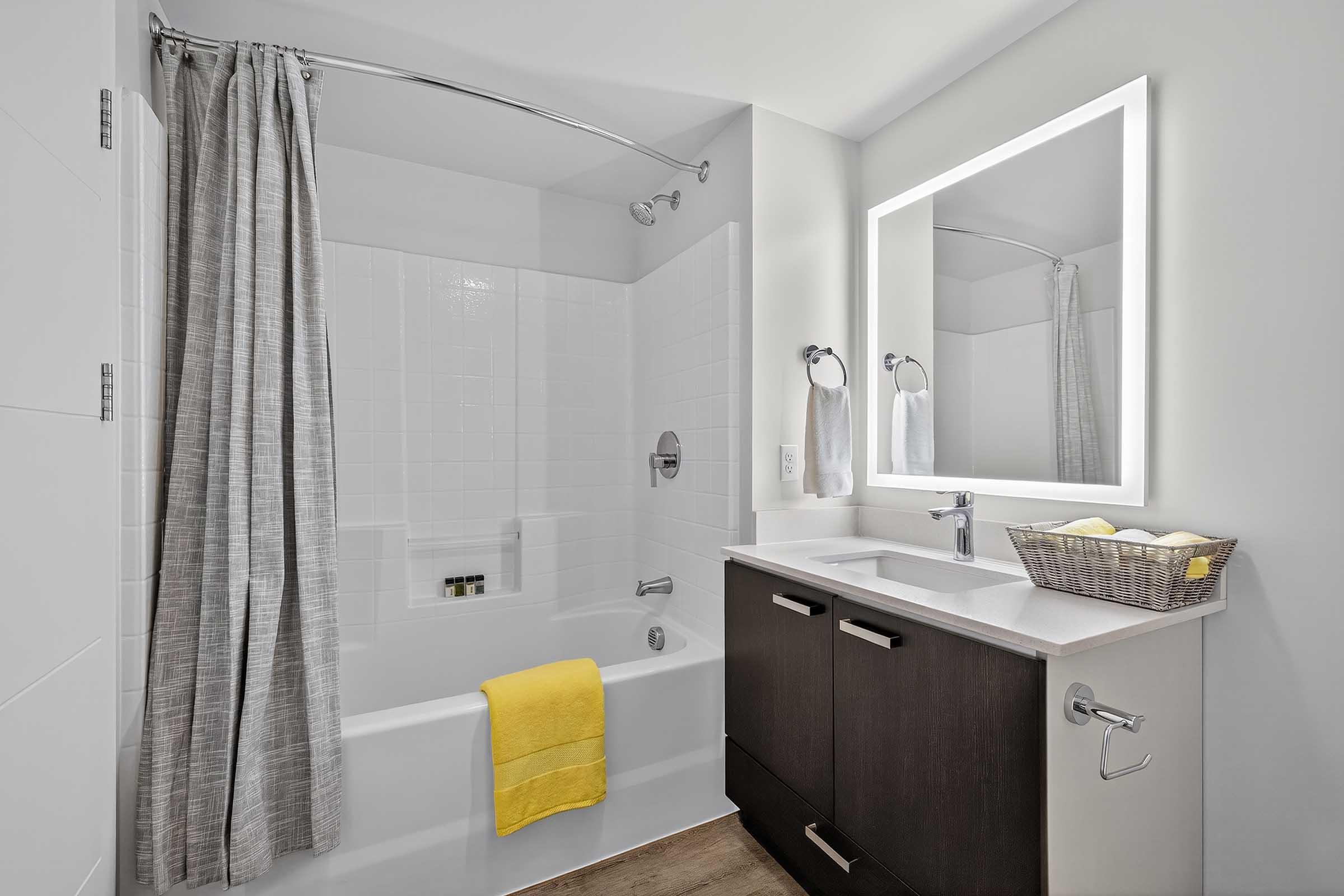 Modern bathroom featuring a white tiled shower with a bath, a gray curtain, a sleek vanity with dark wood cabinetry, and a large illuminated mirror. A yellow towel is placed on the tub, while a basket with towels sits on the countertop. Good lighting creates a bright and clean atmosphere.