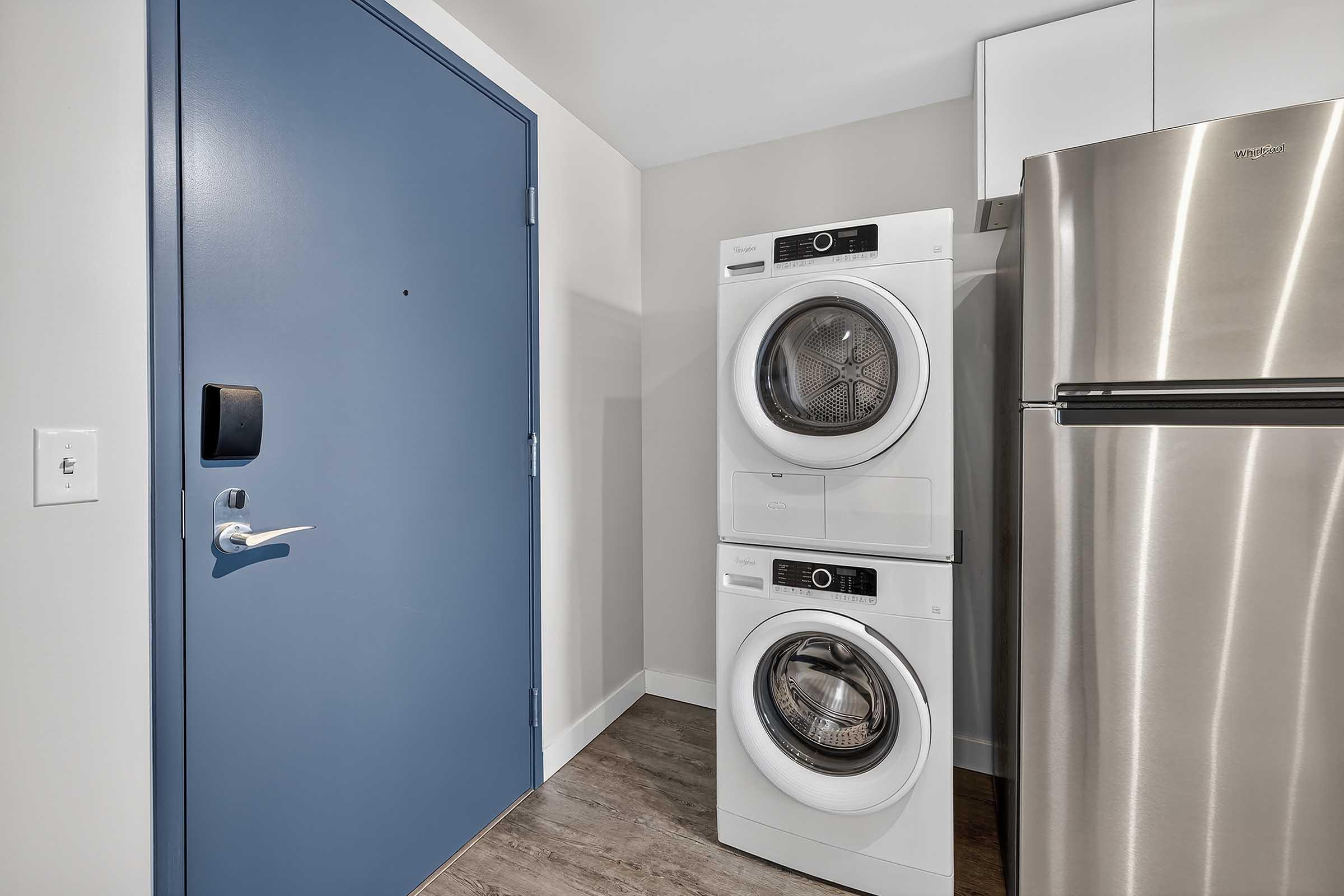 Stacked washer and dryer units next to a stainless steel refrigerator in a modern laundry area. A blue door is visible in the background, adding a pop of color to the otherwise neutral-toned space. The flooring appears to be wood-like laminate.
