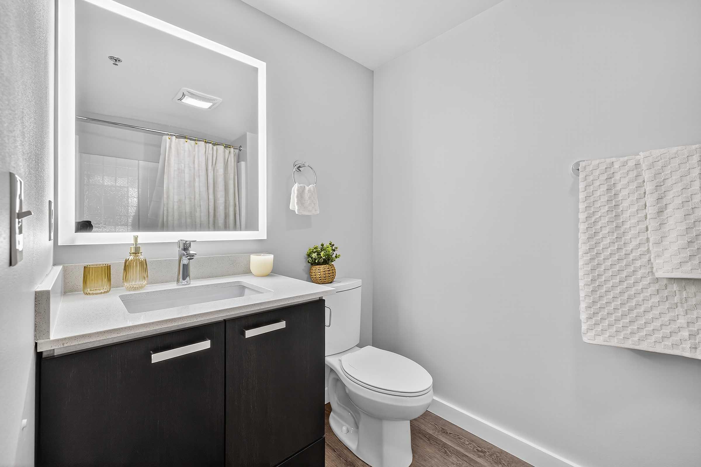 A modern bathroom featuring a white and gray color scheme. It includes a sleek countertop with a sink, a mirror with soft backlighting, a shower curtain in the background, a decorative plant, and neatly arranged towels on a rack. The floor is made of light wood, contributing to a clean and contemporary look.