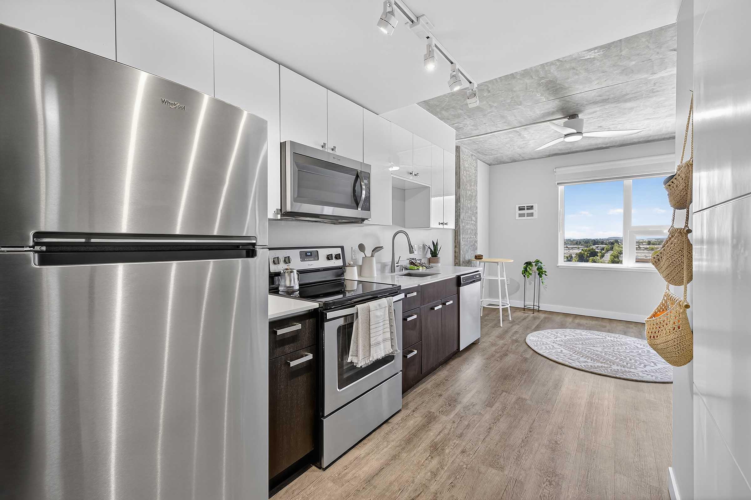 Modern kitchen featuring stainless steel appliances, including a refrigerator and oven. Dark wooden cabinets complement the light countertops. A window offers a view of the outside, while a round rug and a small plant add a touch of decor. Bright and airy atmosphere with natural light.