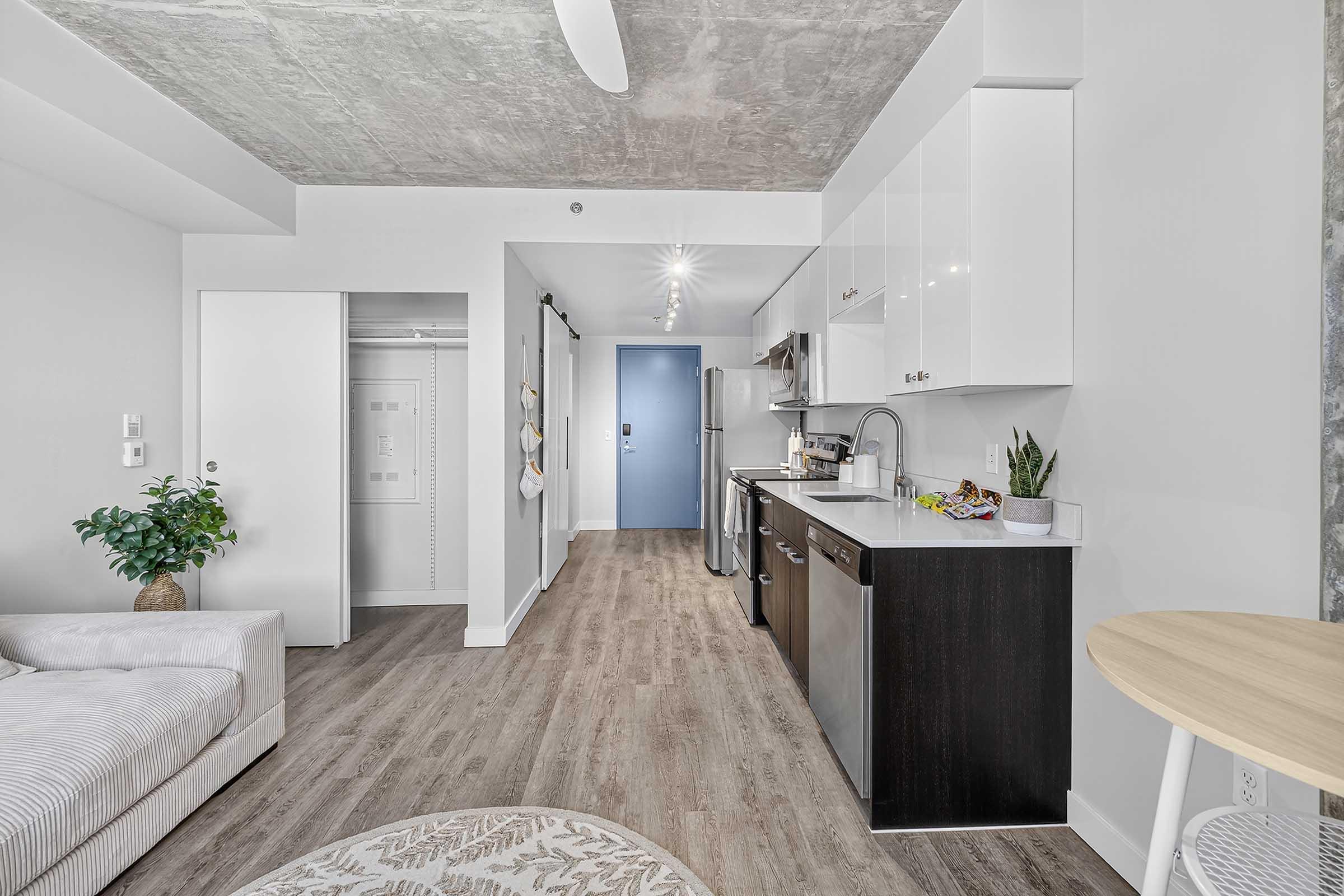 A modern kitchen area with white cabinets and stainless steel appliances, including a fridge and stove. There's a small dining table, a light-colored sofa, and a decorative plant. The floor is wooden, and the walls are light-colored, creating a bright and airy atmosphere.