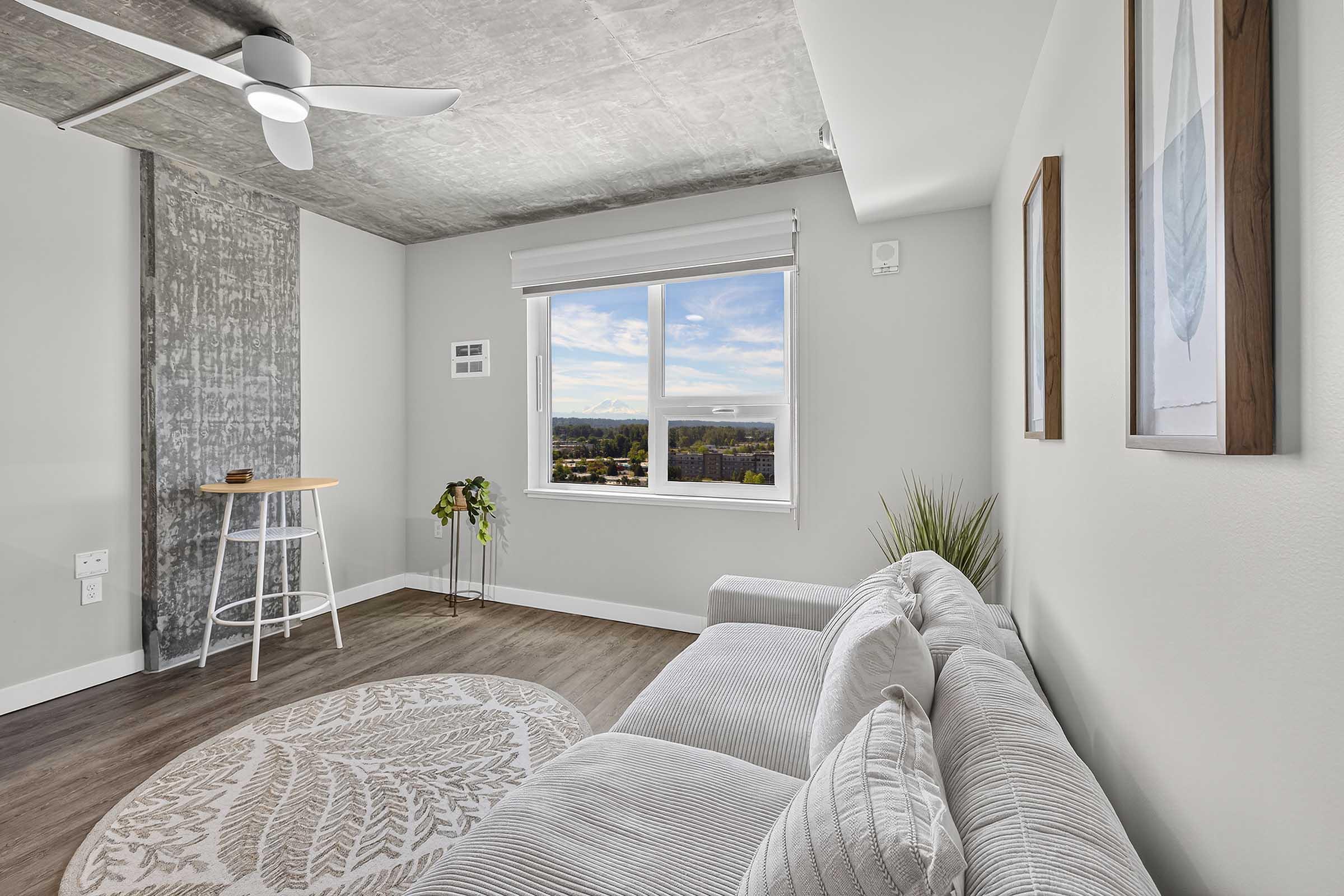 A modern living room featuring a light gray sofa with textured cushions, a round decorative rug on wooden flooring, and a small white table with a barstool. Large window offers a view of the landscape outside, and the walls are painted in soft tones with minimalist wall art and a potted plant adding a touch of greenery.