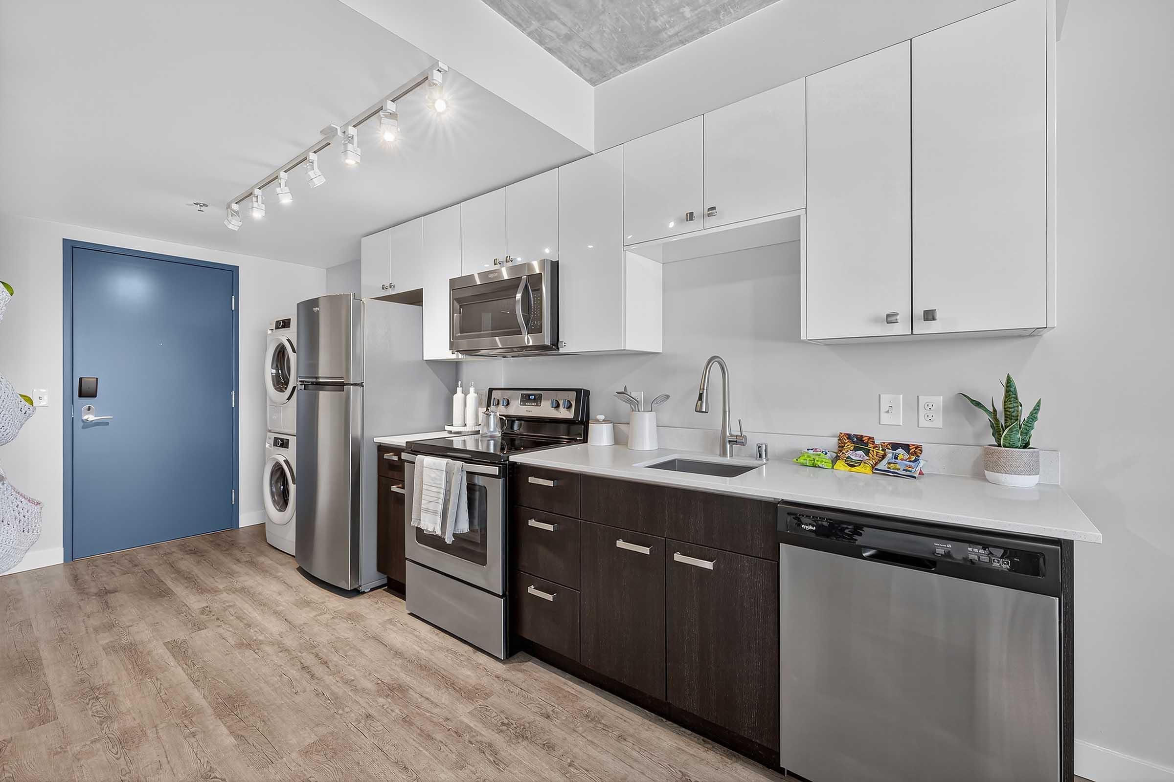 Modern kitchen featuring stainless steel appliances, including a refrigerator, oven, and dishwasher, with sleek white cabinetry and dark wood accents. The countertop is light-colored with a sink and decorative plants. A laundry area with stacked washer and dryer is visible, along with a blue door.