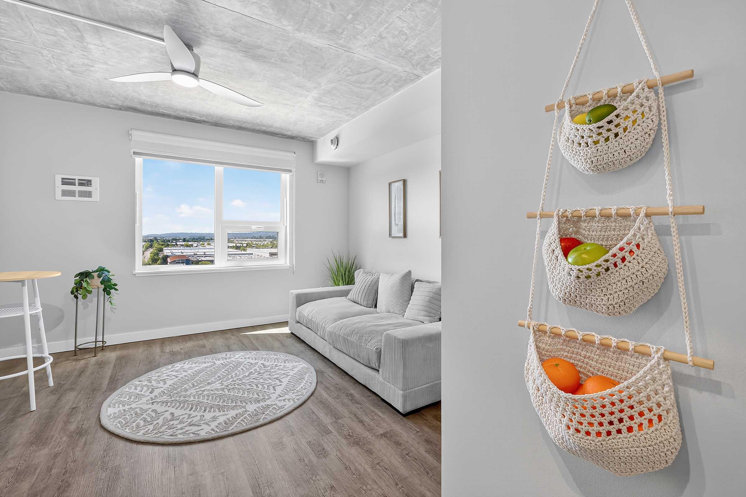 A modern, bright living room featuring a gray sofa, a round patterned rug, and a large window with a view. On the wall, there are three hanging woven baskets containing colorful fruit. A ceiling fan and potted plant add to the contemporary decor.