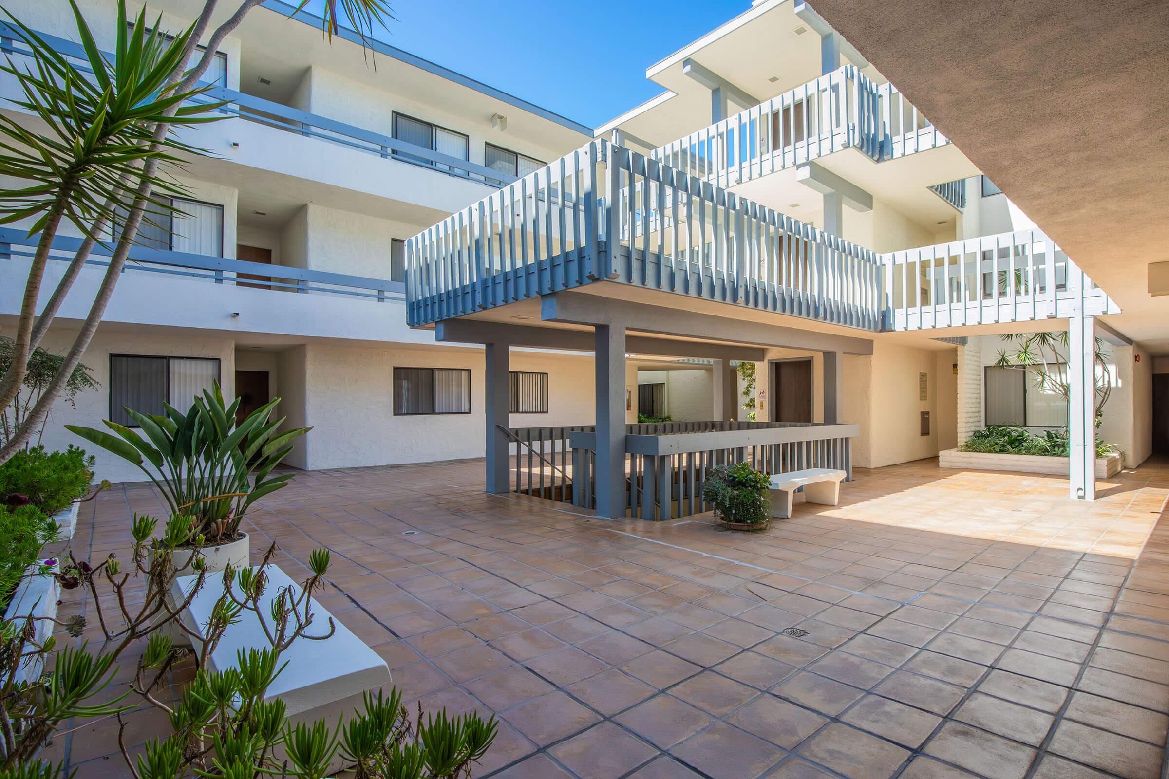 A view of a courtyard in an apartment complex featuring two levels of balconies, surrounded by greenery. The tiled ground showcases a spacious area with plants, offering a bright and open atmosphere under a clear blue sky.