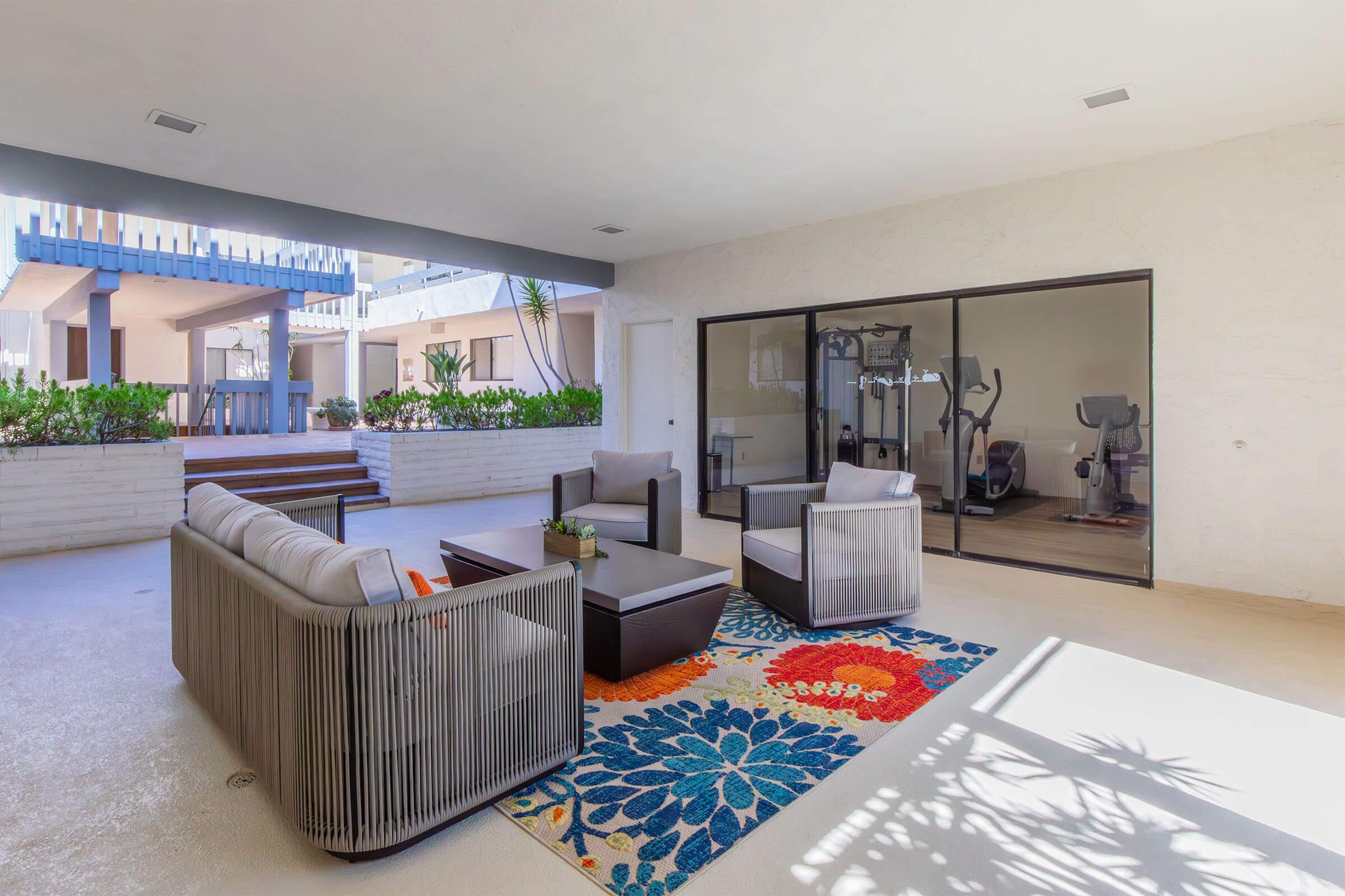 A modern outdoor living area featuring a cozy seating arrangement with gray sofas and a dark coffee table, complemented by a vibrant floral area rug. Large sliding glass doors provide access to an interior space, while greenery and sunlight create a bright, inviting atmosphere.