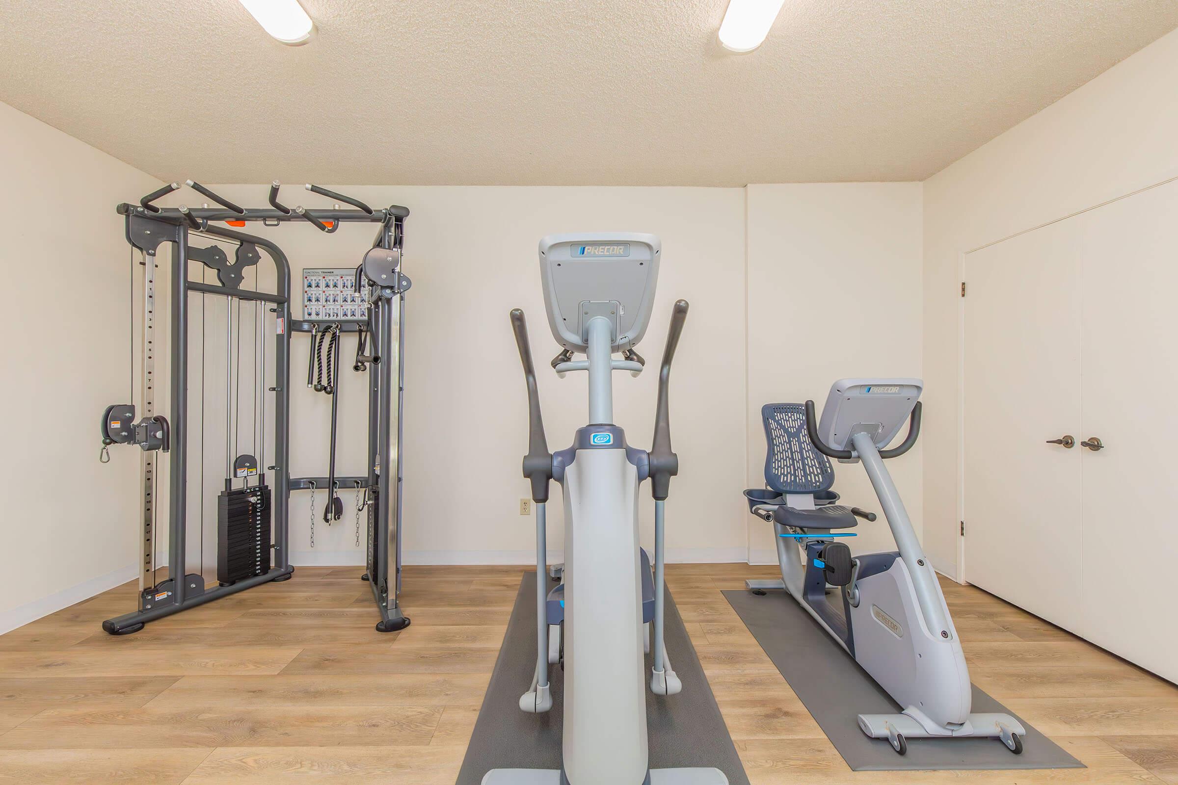 A bright and spacious home gym featuring a multi-station strength training machine, an elliptical machine, and a recumbent bike on wooden flooring. The walls are painted light, and the room is well-lit with overhead lighting.