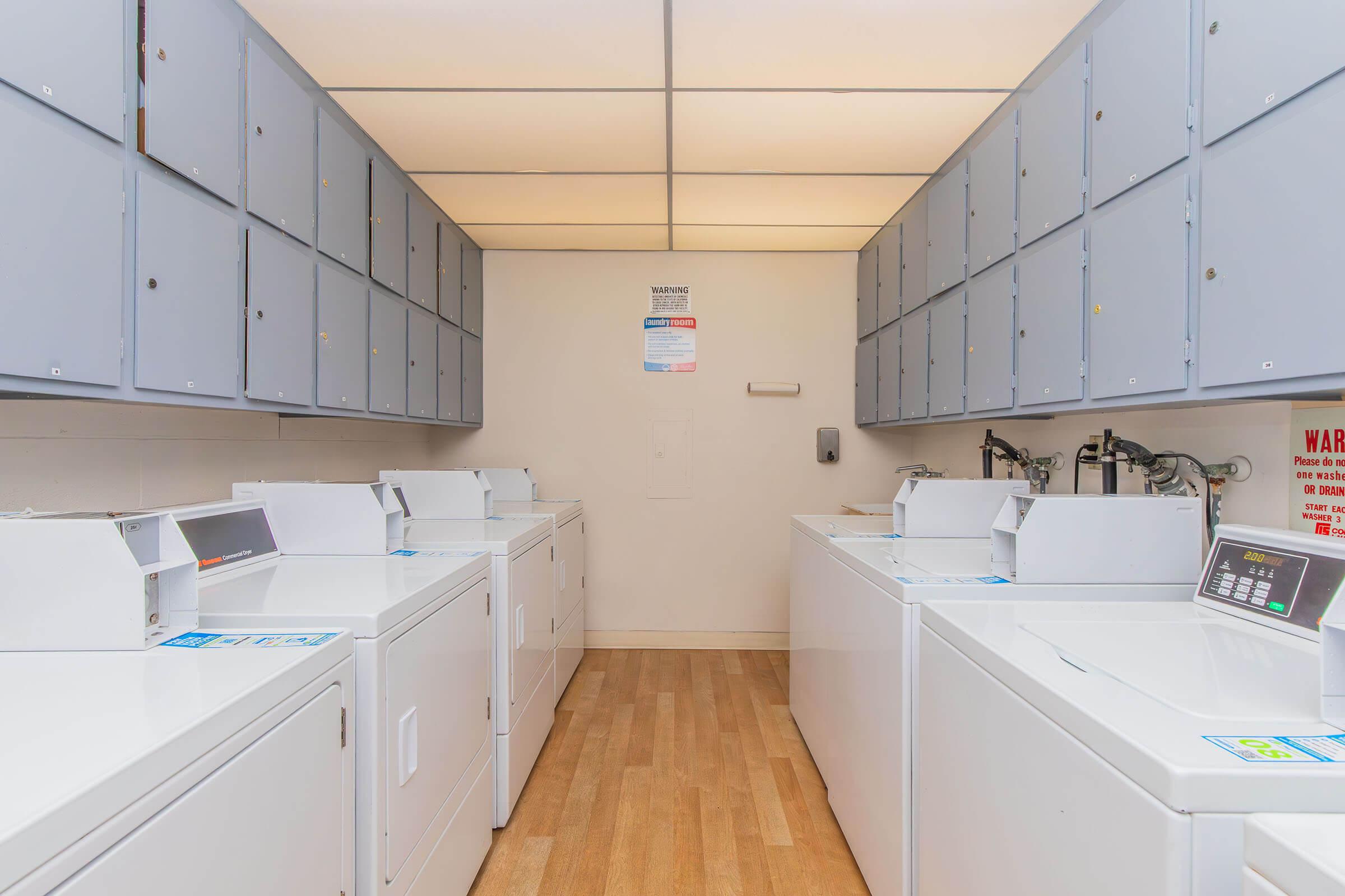 A clean, bright laundry room featuring several stacked white washing machines and dryers. The machines are neatly arranged in two rows, with gray storage cabinets above. The floor is wooden, and the ceiling is well-lit. A warning sign is visible on the wall.