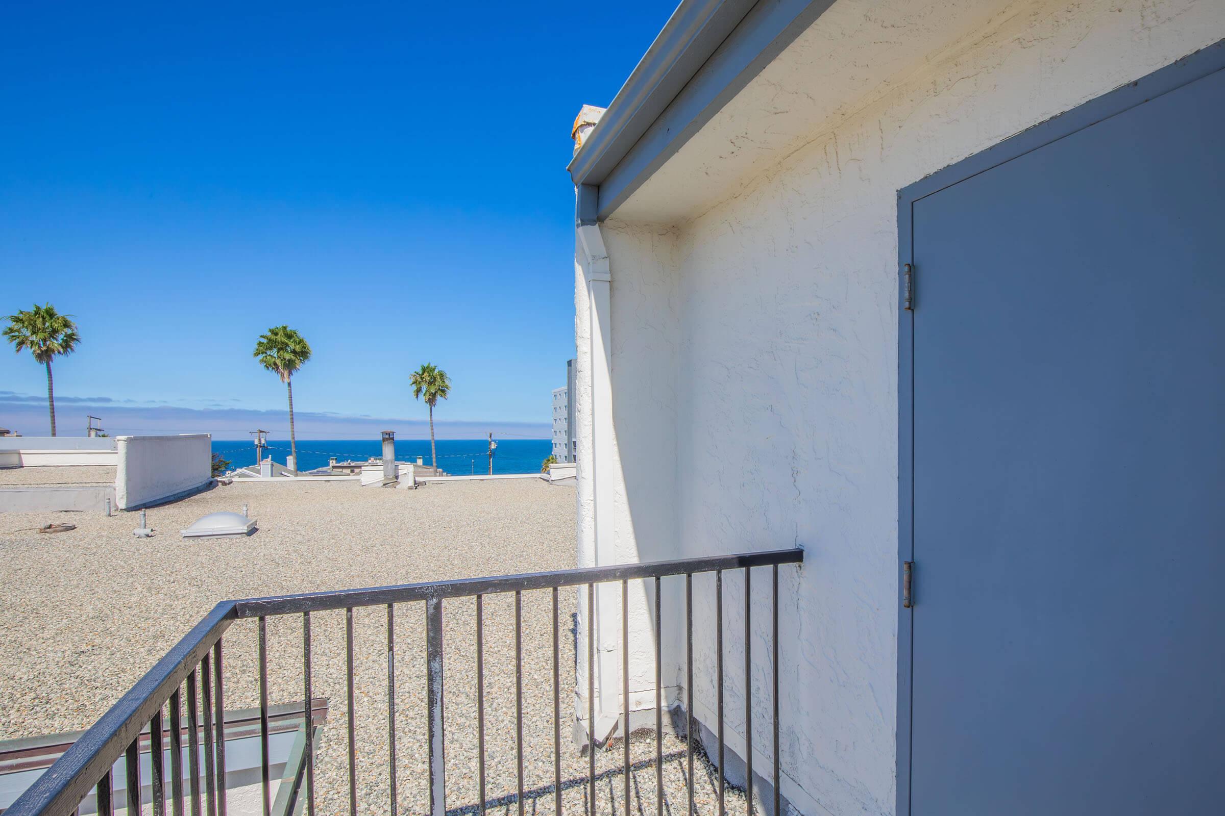 A bright blue sky above a rooftop with palm trees in the background. A door is seen on a white wall, and the rooftop surface is covered with gravel. The ocean is visible in the distance, creating a serene coastal view.