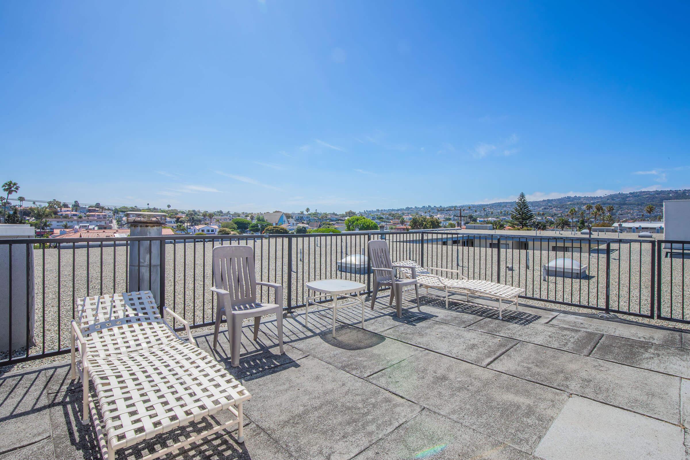 A rooftop patio featuring several white lounge chairs on a sunny day. In the background, there are residential buildings, trees, and a clear blue sky with a few clouds, creating a relaxing outdoor atmosphere.