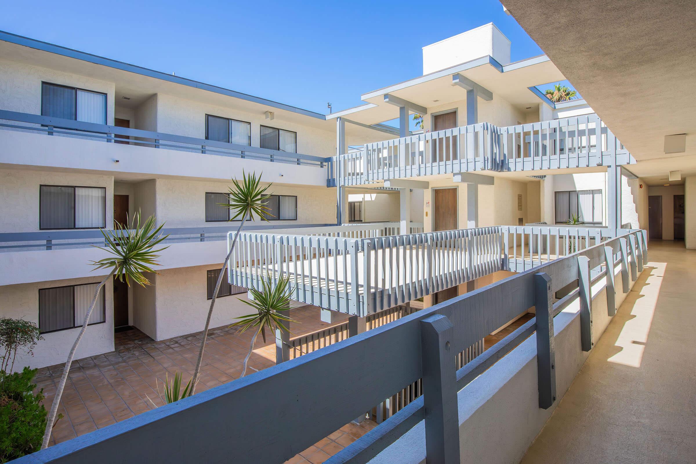 A modern, multi-level apartment complex featuring a central courtyard. The image shows a balcony walkway with railings, surrounded by palm trees and blue skies. The architecture is contemporary with white walls and large windows, creating a bright and inviting atmosphere.