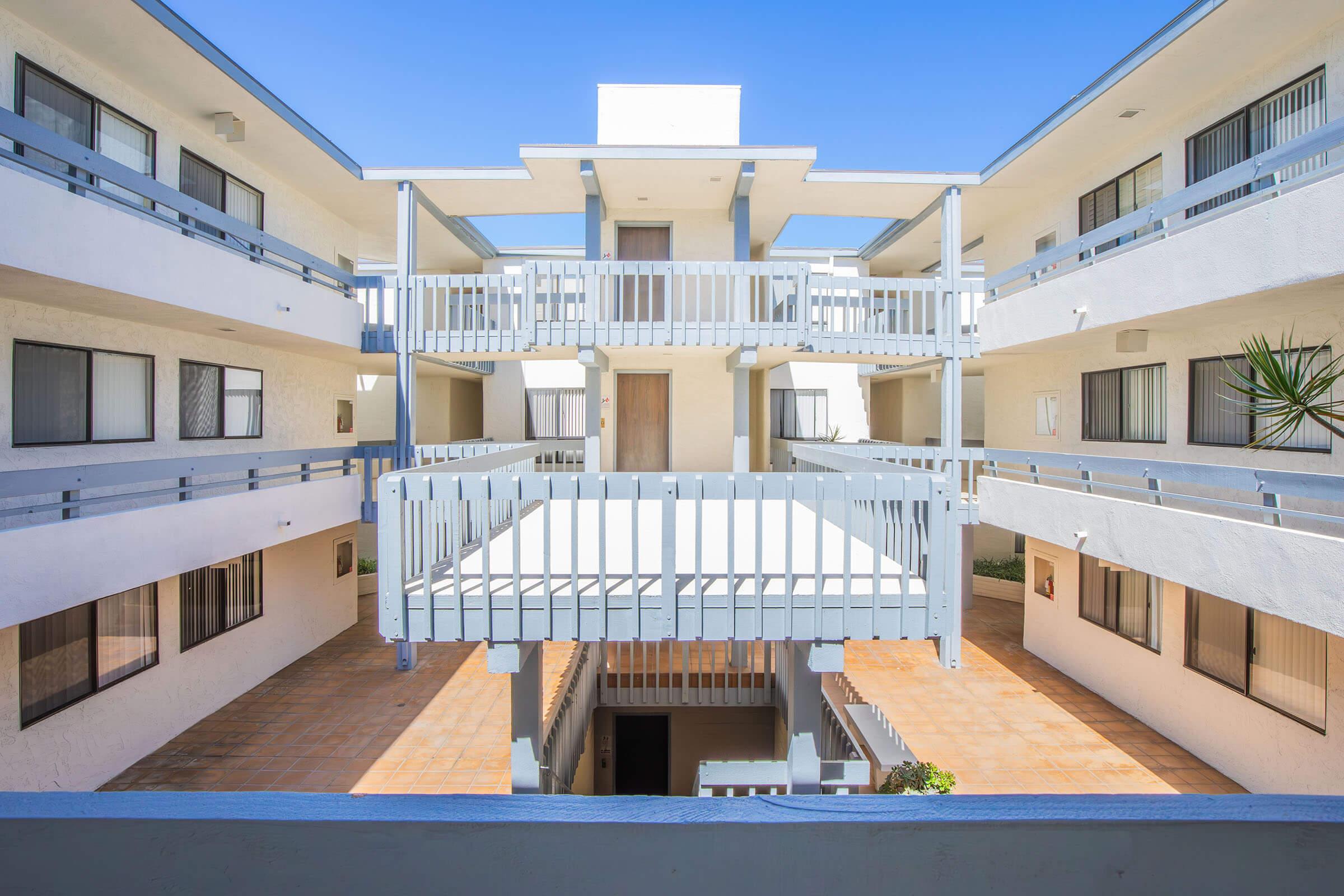 View of a modern courtyard in a two-story apartment building. Features include multiple balconies with blue railings, white exterior walls, and clear blue skies overhead. The central area is paved with terracotta tiles, surrounded by walls that have large windows and doors.