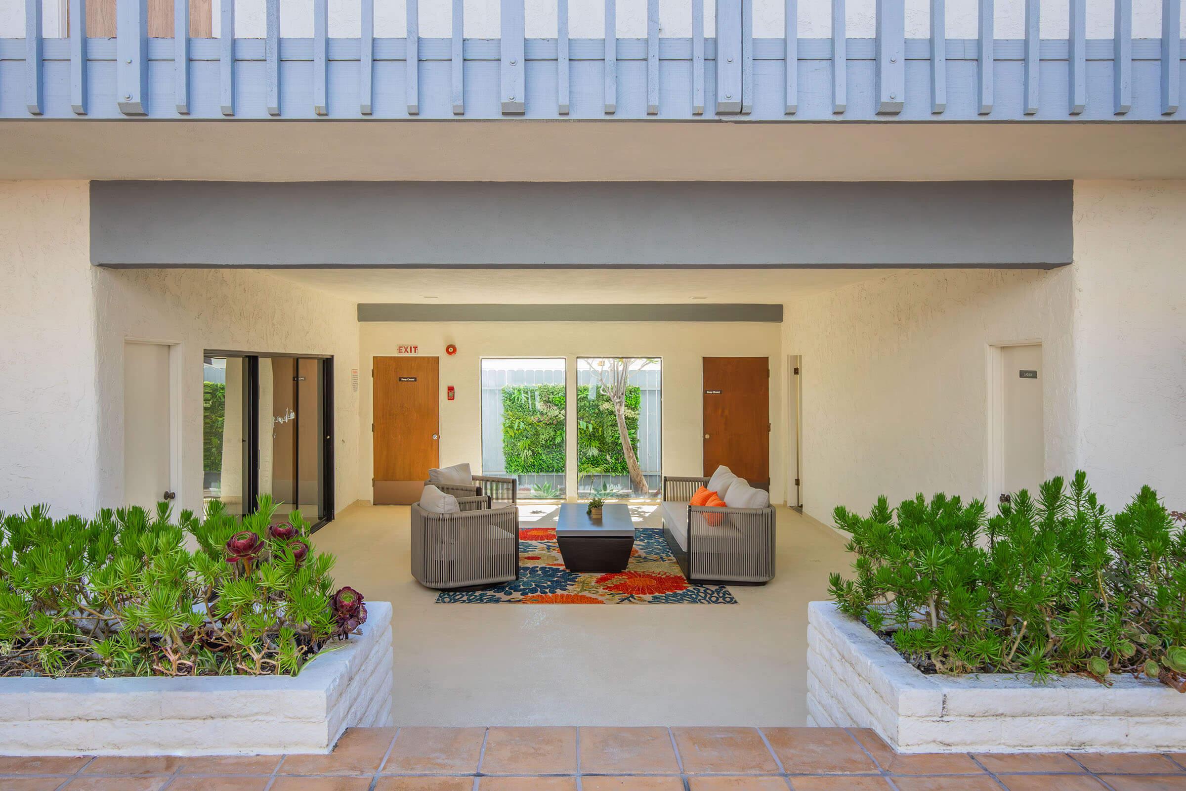 A cozy outdoor seating area featuring two light gray armchairs and a round black coffee table on a colorful rug. Surrounding the space are green plants and a pathway leading to doors. The backdrop includes a wall with a balcony and a view of greenery beyond.