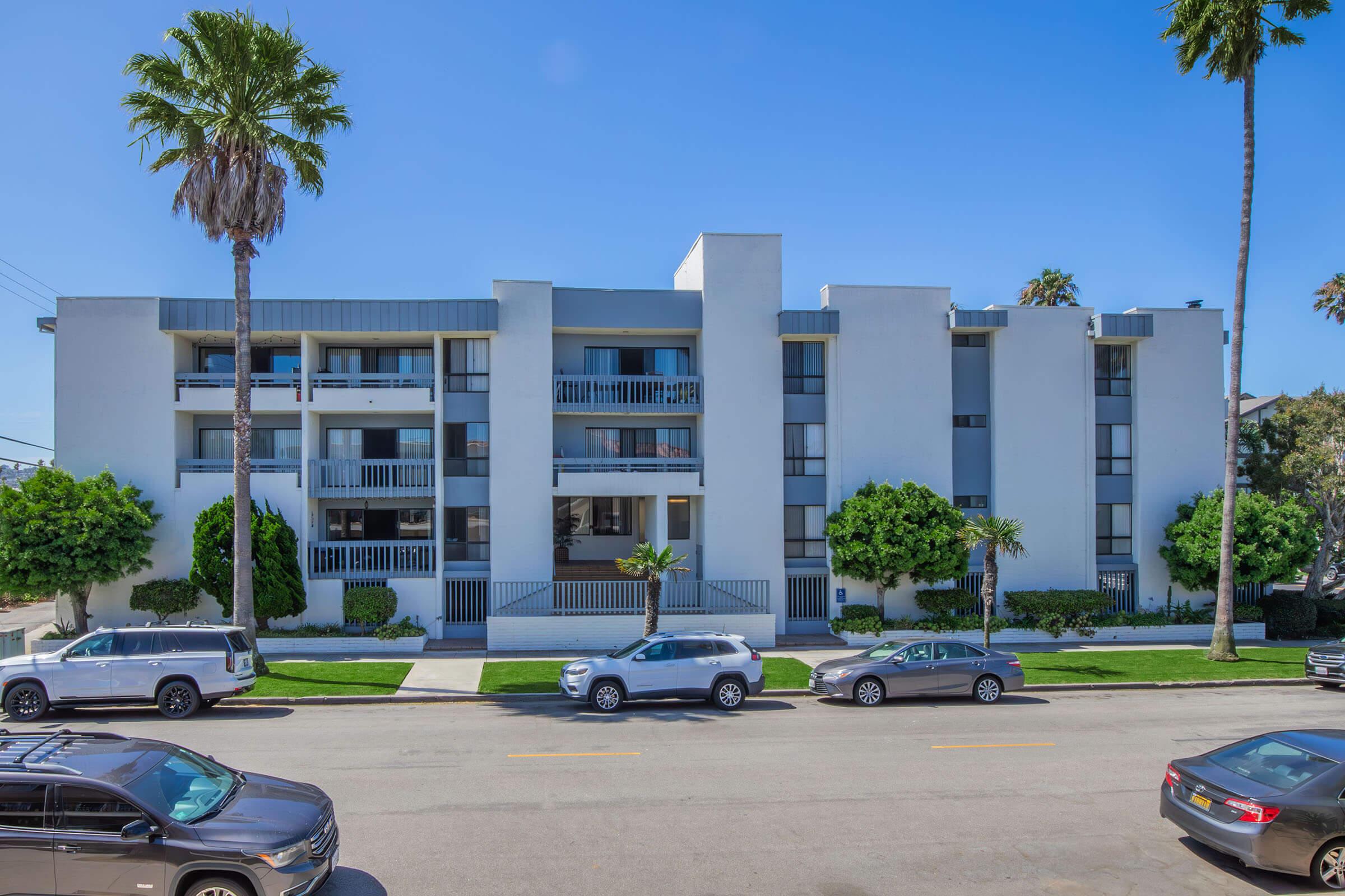 A modern, three-story apartment building with a clean white facade. It features balconies on each floor, surrounded by palm trees and well-maintained greenery. In the foreground, parked cars line the street under a clear blue sky. The building is set in a sunny, urban neighborhood.