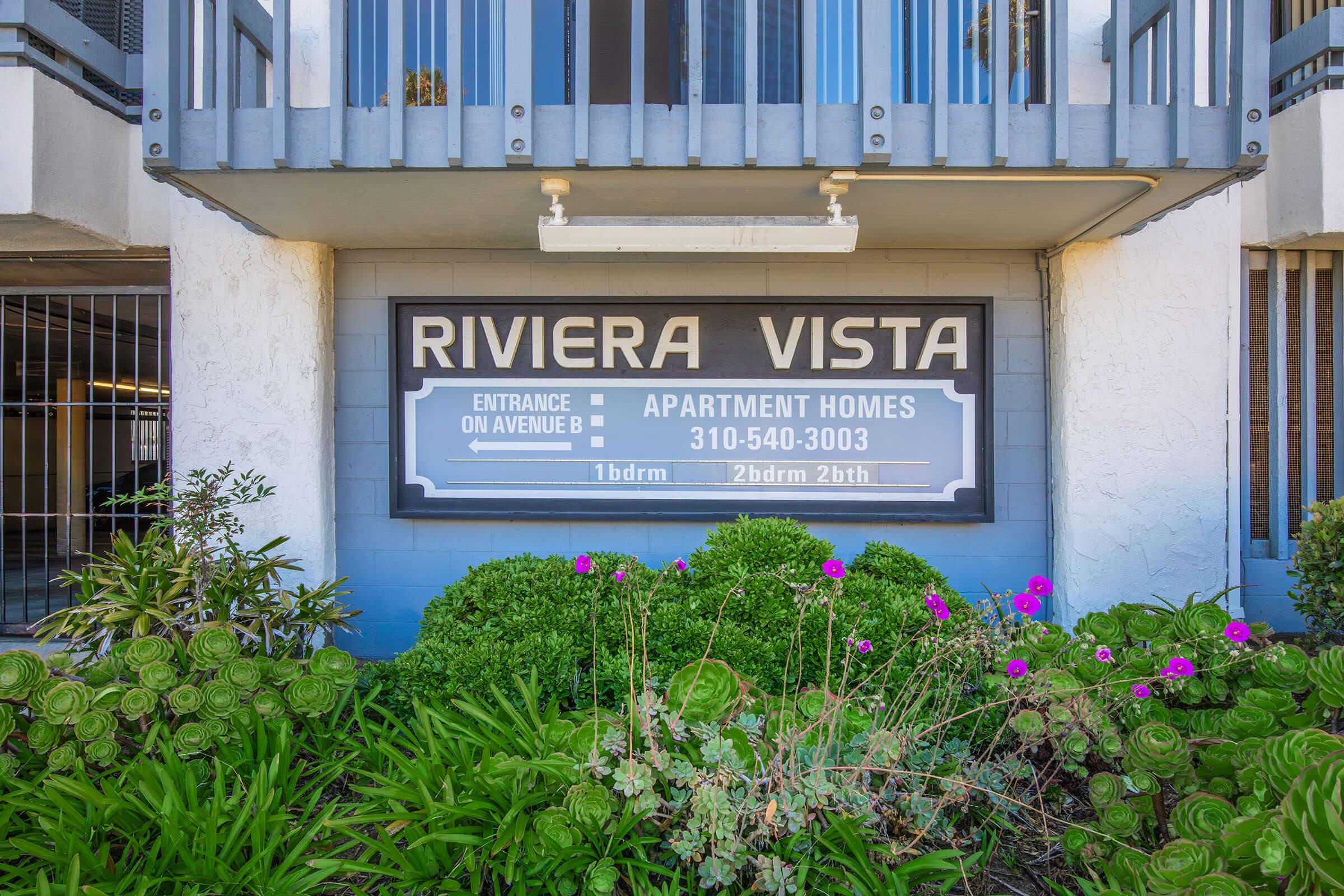 Sign for "Riviera Vista" apartment homes displaying contact number 310-540-3003, featuring details for 1-bedroom and 2-bedroom units. The sign is surrounded by greenery and pink flowers, positioned at the entrance on Avenue J.