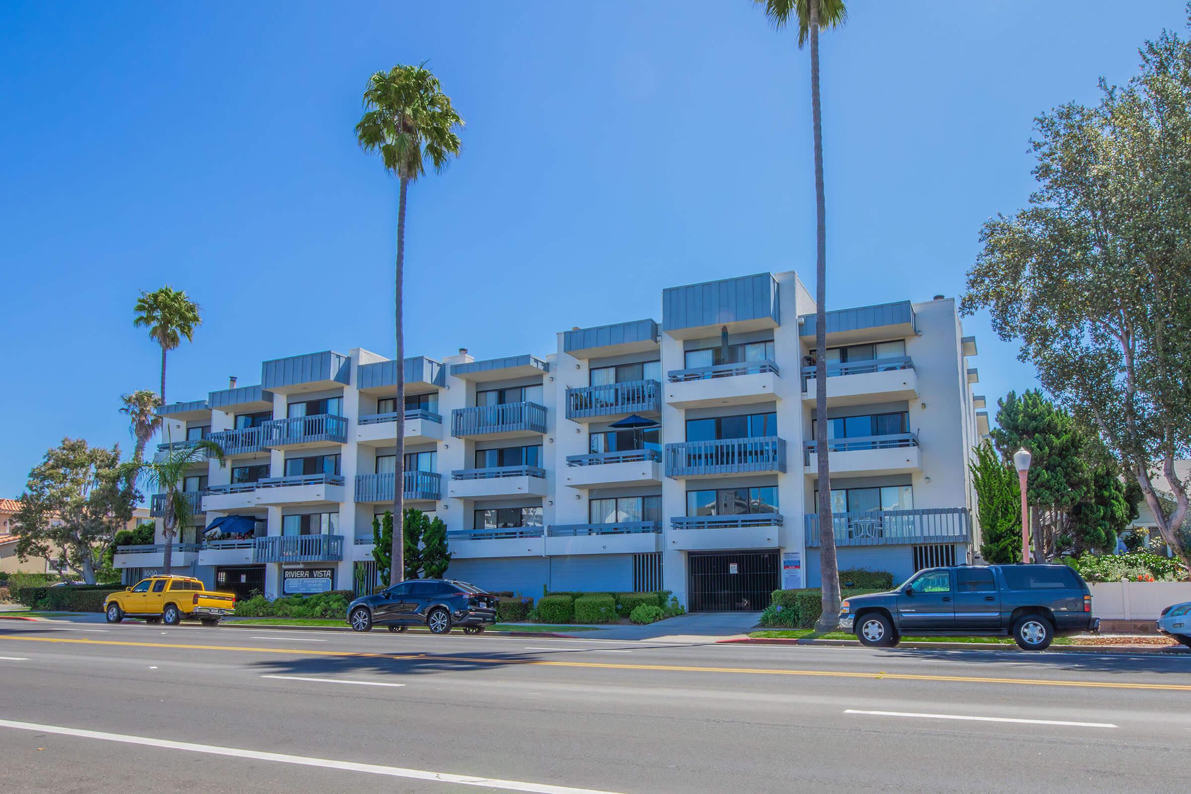 Modern multi-story residential building with balconies, flanked by tall palm trees. A yellow taxi and a black SUV are parked in front. The sky is clear and blue, creating a bright and sunny atmosphere. Sidewalk and road visible in the foreground.