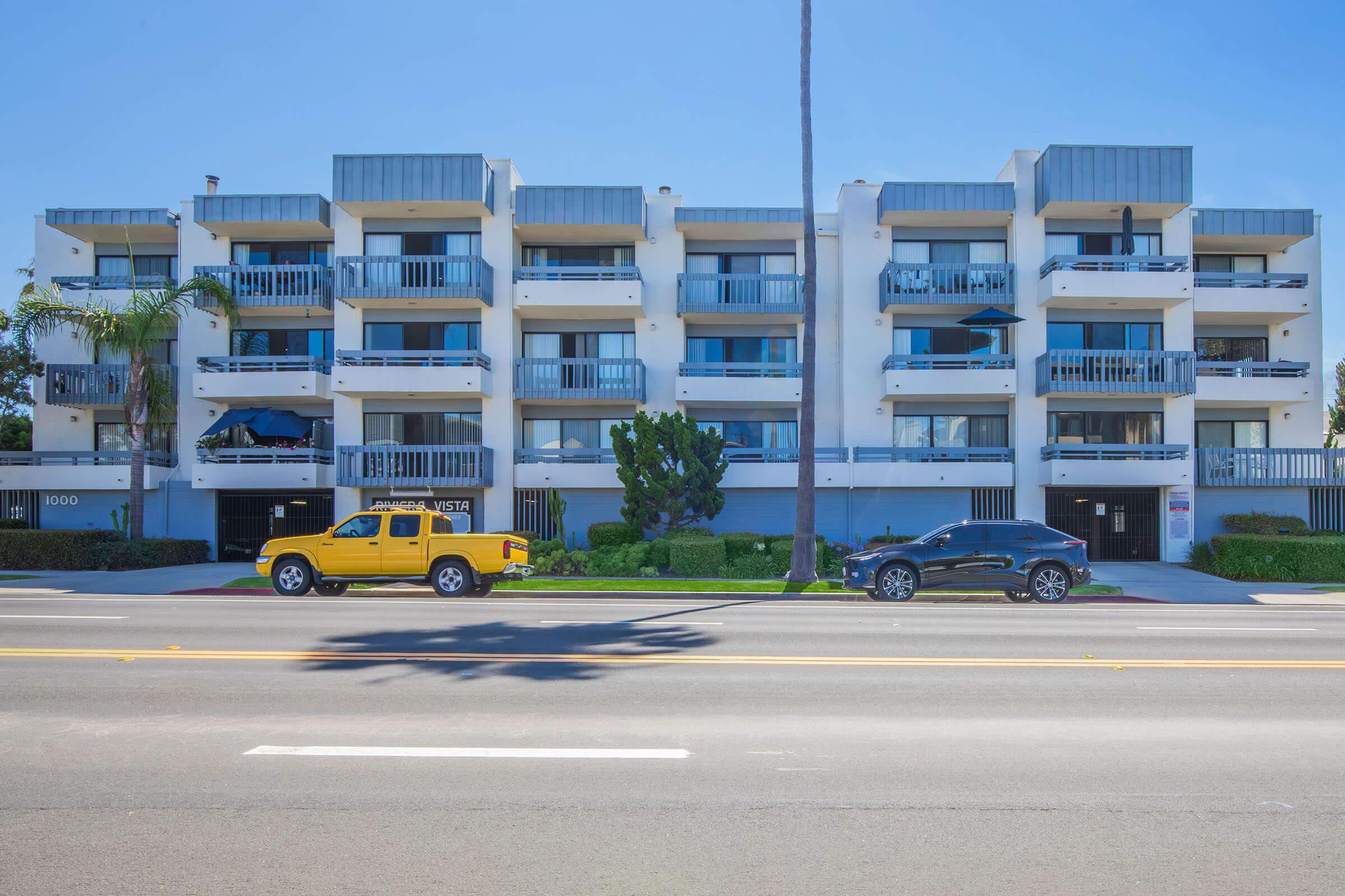 A modern multi-story building with balconies, situated along a street lined with palm trees. Two vehicles are parked in front: a bright yellow pickup truck and a black car. The sky is clear and blue, creating a vibrant atmosphere. The building features a clean, contemporary design with white walls and gray accents.