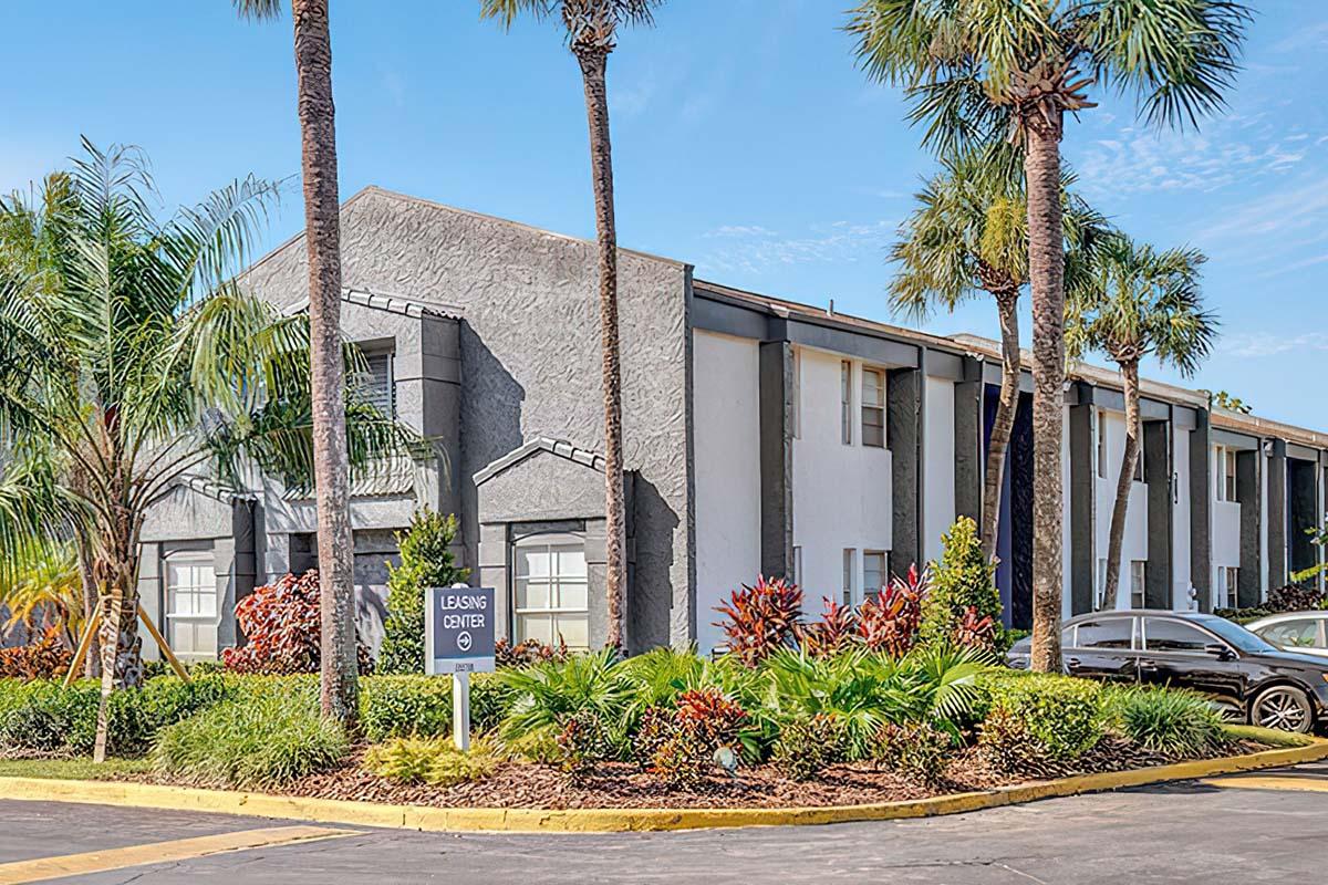 A modern leasing center building with a gray exterior surrounded by lush tropical landscaping, including palm trees and colorful plants. The entrance features a sign indicating it is the leasing center, with a clear blue sky in the background.