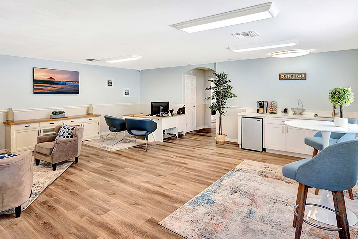 A modern office space featuring two desks with computers, a seating area with armchairs, and decorative elements like plants and artwork. Light wood flooring and bright lighting create an inviting atmosphere. A coffee bar area is visible, adding to the workspace's comfort.