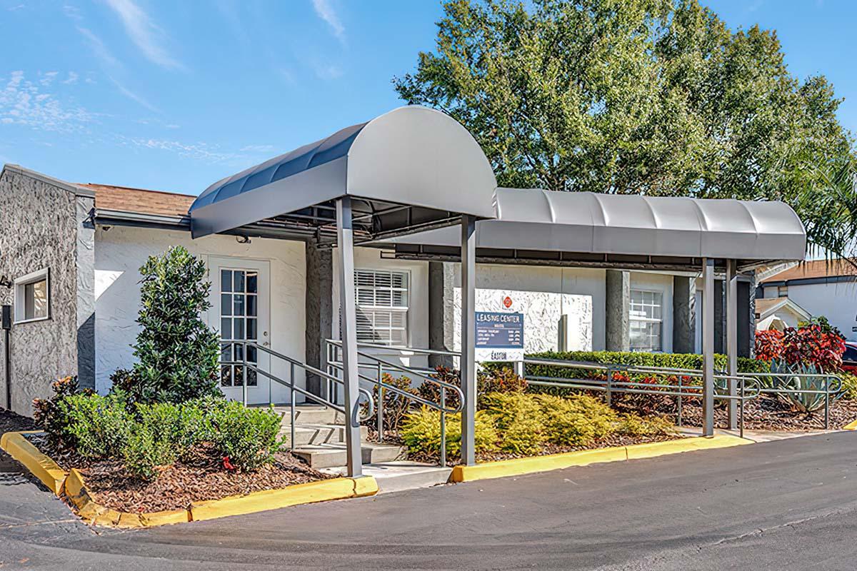 A single-story building with a covered entrance and ramp, surrounded by landscaped greenery and colorful shrubs. The structure features a light-colored exterior and is situated in a paved parking area under a blue sky.